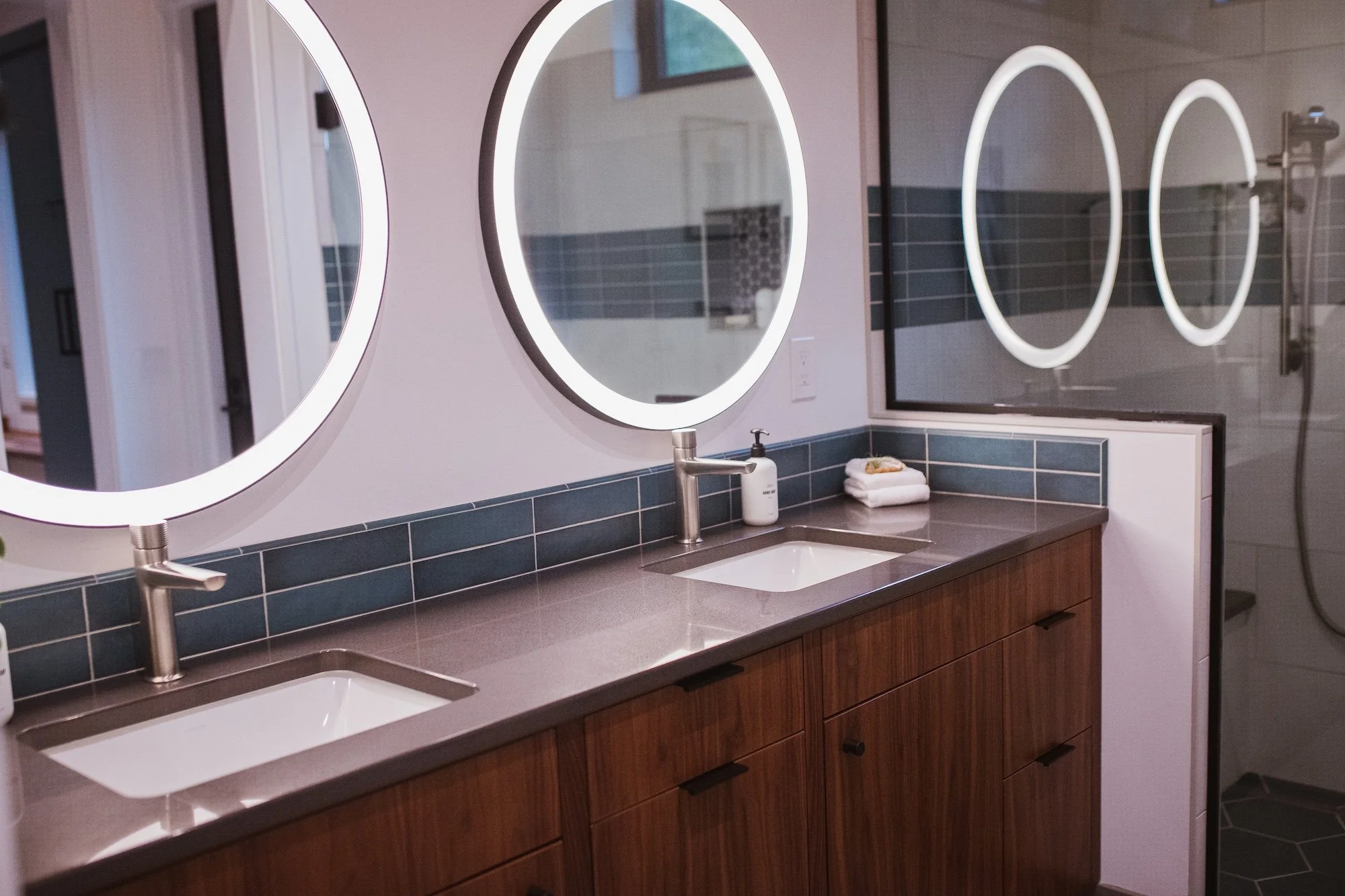 Public restroom with three illuminated oval mirrors above a double sink vanity with wooden cabinets and black tiled backsplash, soap dispensers, and folded towels on the counter. A shower area with a glass door and showerhead is visible in the reflec