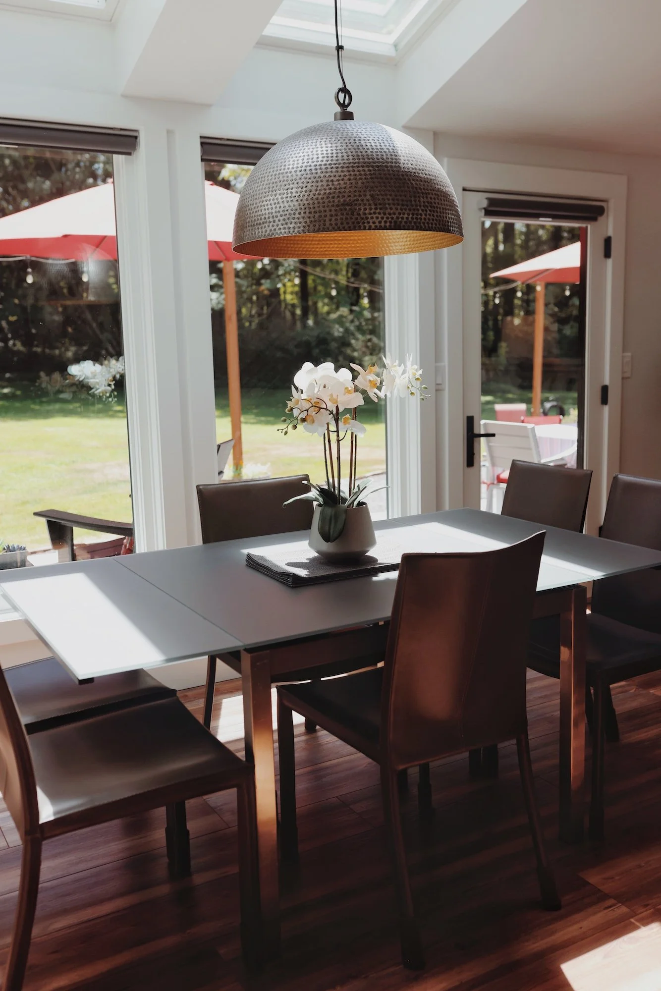 A dining room with a white table, surrounded by dark wooden chairs, a potted white orchid on the table, and large glass doors leading outside to a patio with umbrellas and greenery, lit by sunlight from a skylight.