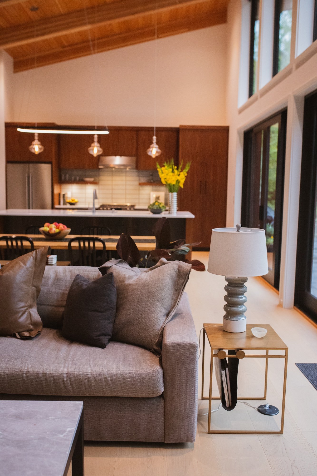 Living room with beige couch, brown cushions, side table with lamp, kitchen in background with black chairs, colorful fruit bowl, yellow flowers, and large windows.