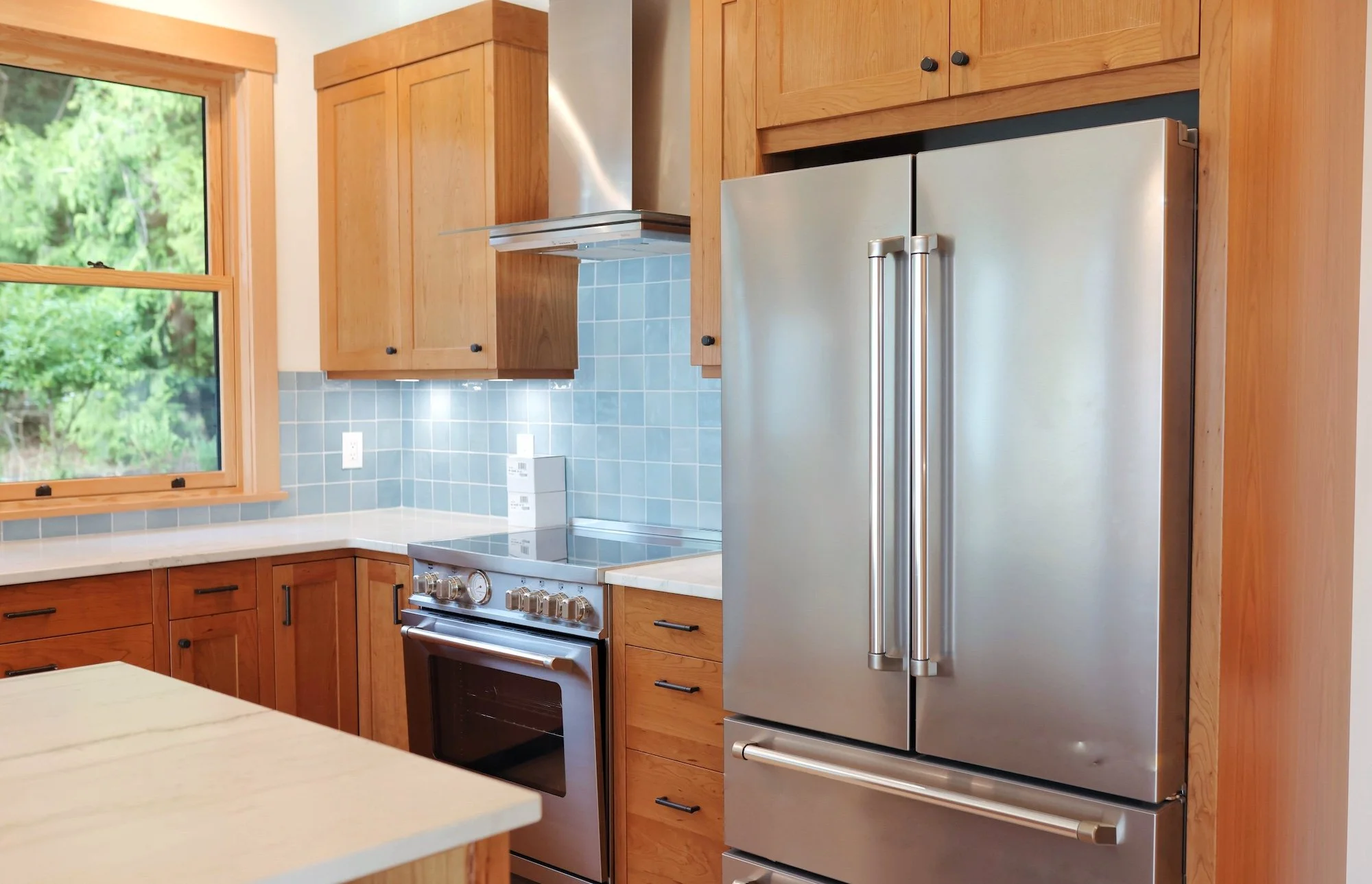 Kitchen with wooden cabinets, a stainless steel refrigerator, a stove, a window showing green trees outside, and blue tiled backsplash.
