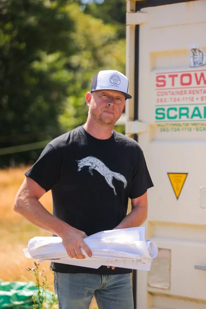 A man wearing a black T-shirt with a white graphic of a jumping animal and a white and black trucker hat stands outdoors holding rolled-up blueprints. Trees and a white rental container are in the background.