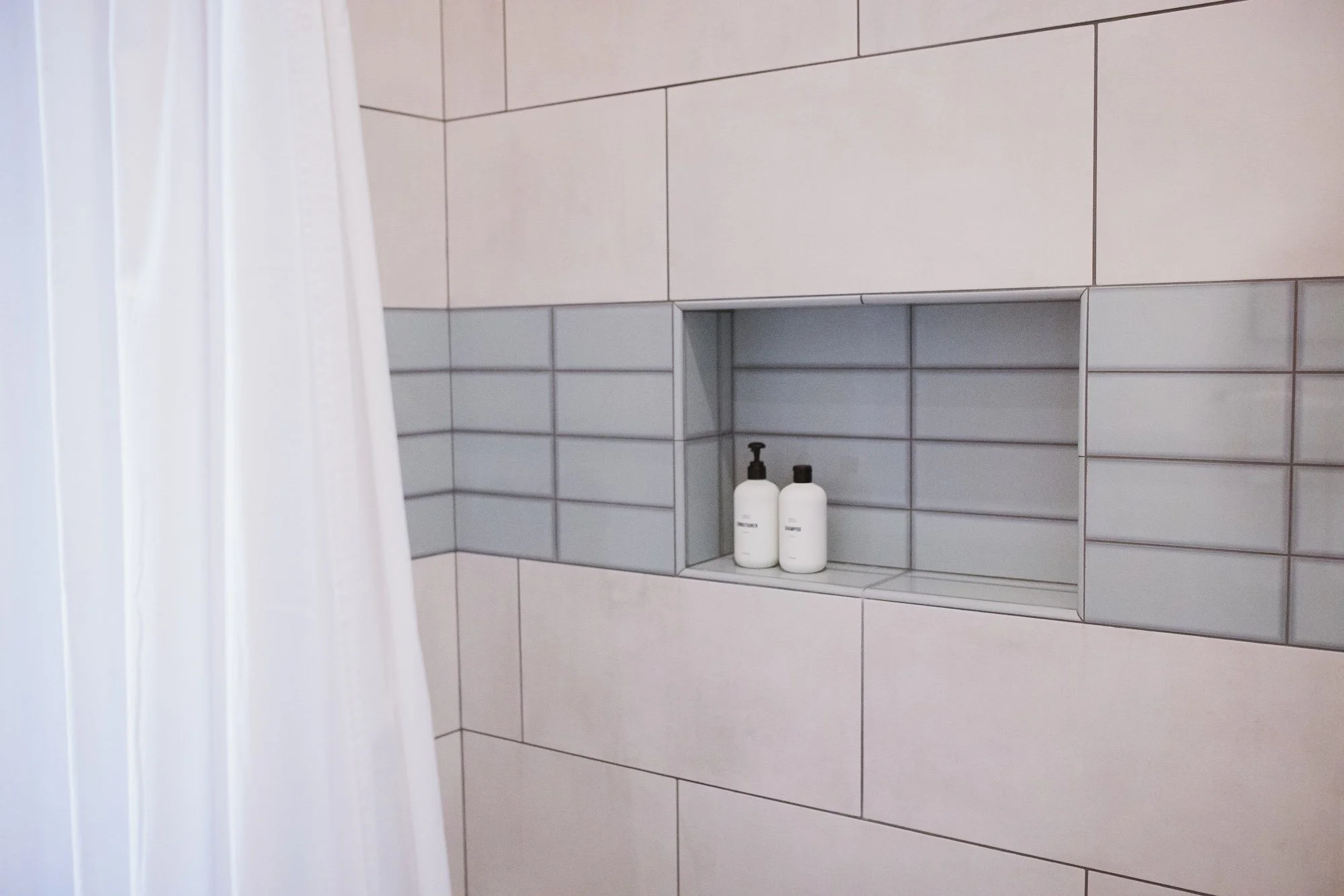 A tiled shower wall with a built-in shelf holding two white bottles, partially obscured white shower curtain on the left.