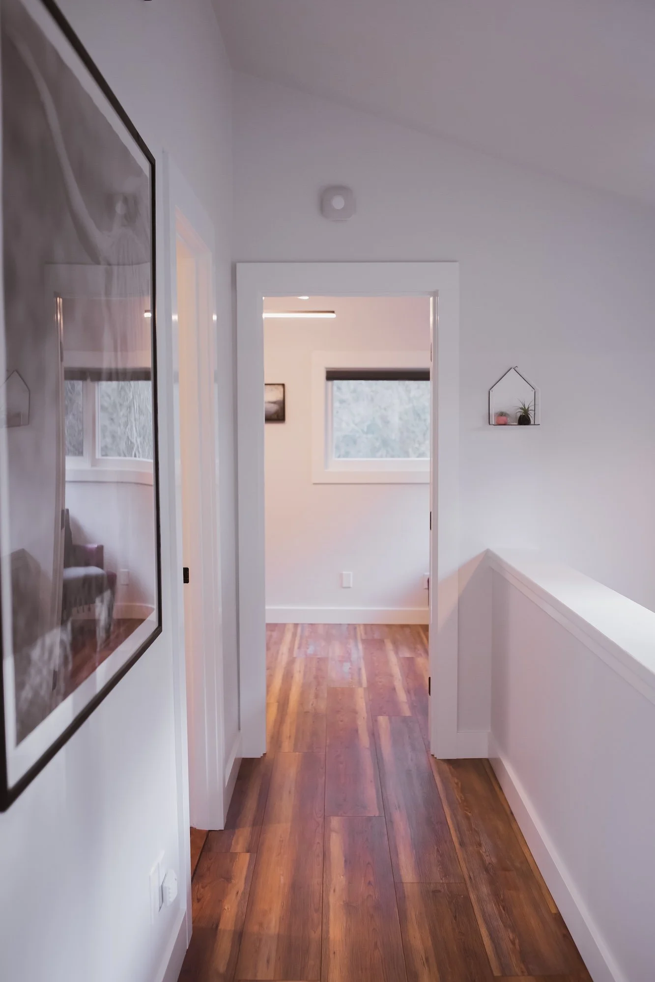 Interior view of a hallway leading to a sunny room with two windows, wooden flooring, and minimalist decor, including small wall shelves with plants.