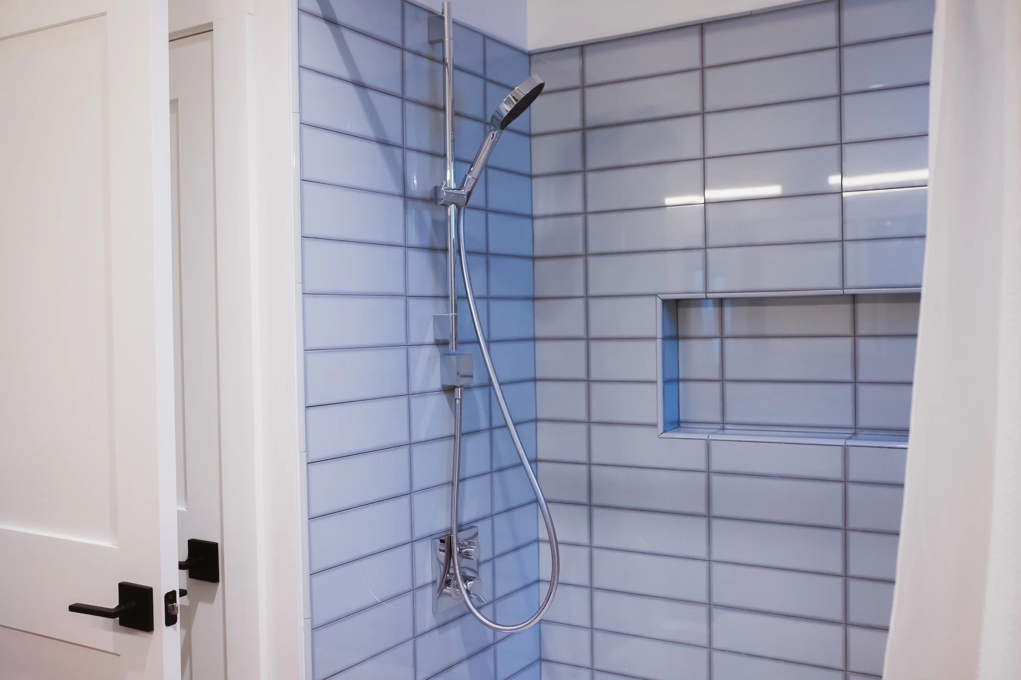 A modern shower area with blue rectangular tile walls, a built-in niche, and a handheld showerhead mounted on a vertical bar.