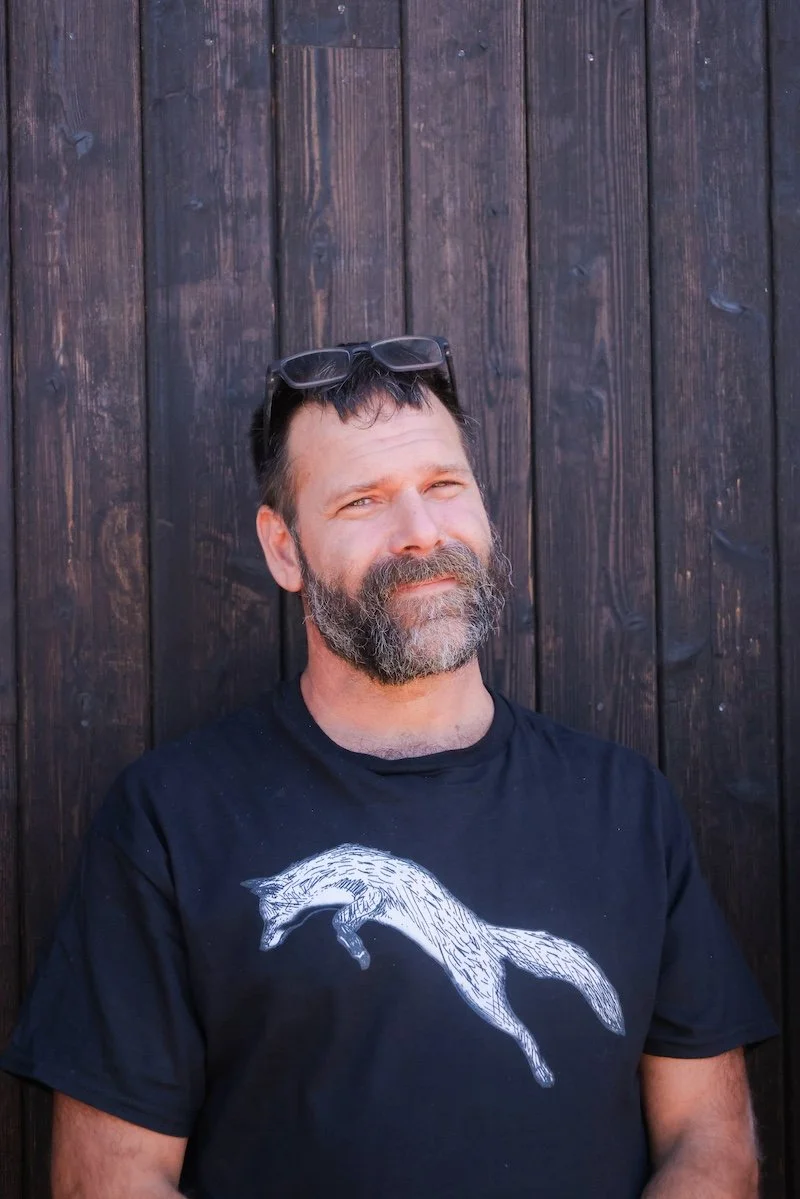A middle-aged man with a beard and short hair, wearing sunglasses on his forehead, standing in front of a dark wooden fence, wearing a black t-shirt with a white graphic of a moving fox.