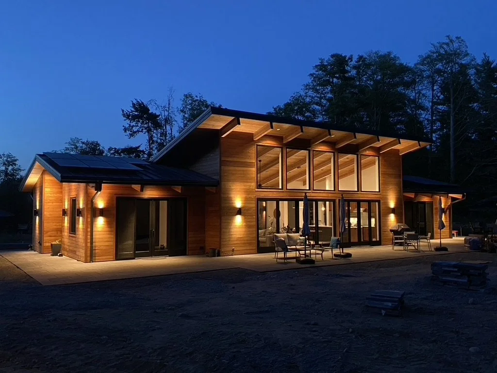 A modern wooden house with large glass windows and sliding doors, lit up at night, surrounded by trees.