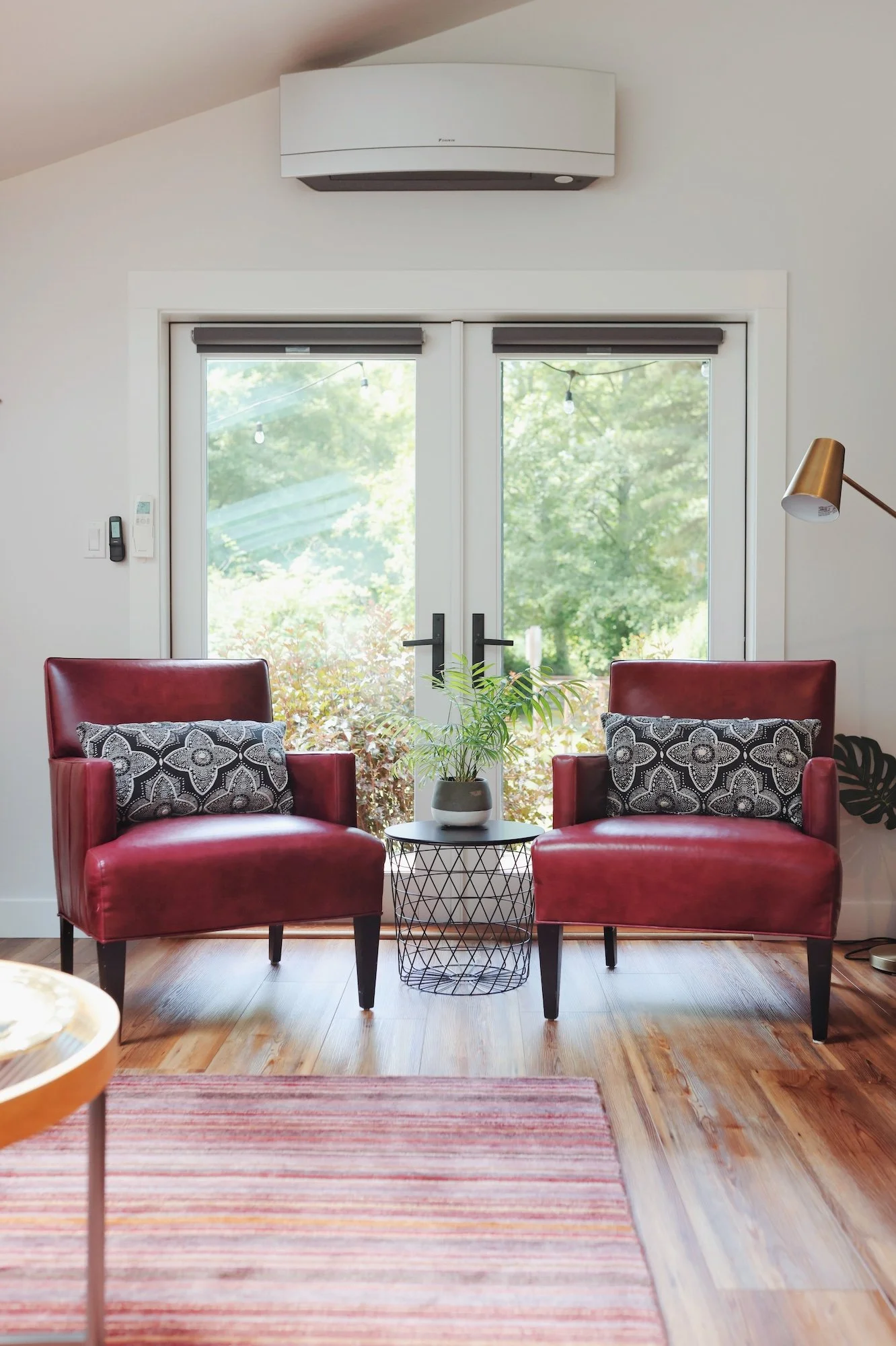 Living room with two red armchairs, a small round table with a potted plant, a striped area rug, sliding glass doors with greenery outside, and a white wall-mounted air conditioner.