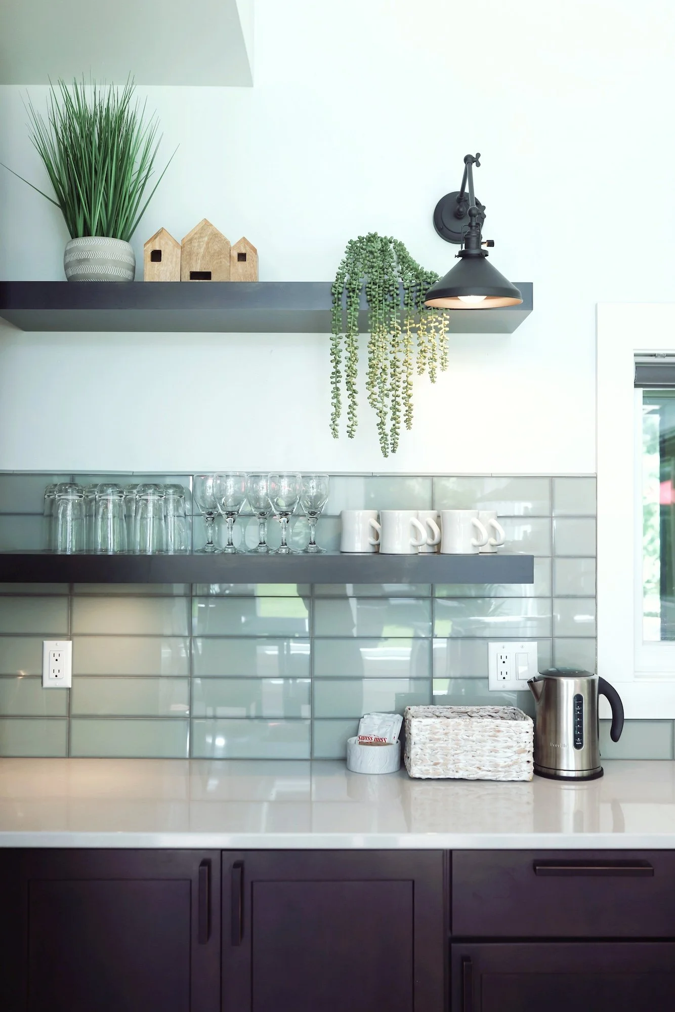 Kitchen with dark cabinets, white countertop, glassware on a gray shelf, white mugs, a soap dish, wicker basket, electric kettle, and a tiled backsplash. There are two shelves with decorative items like a potted plant, small house models, and a hangi