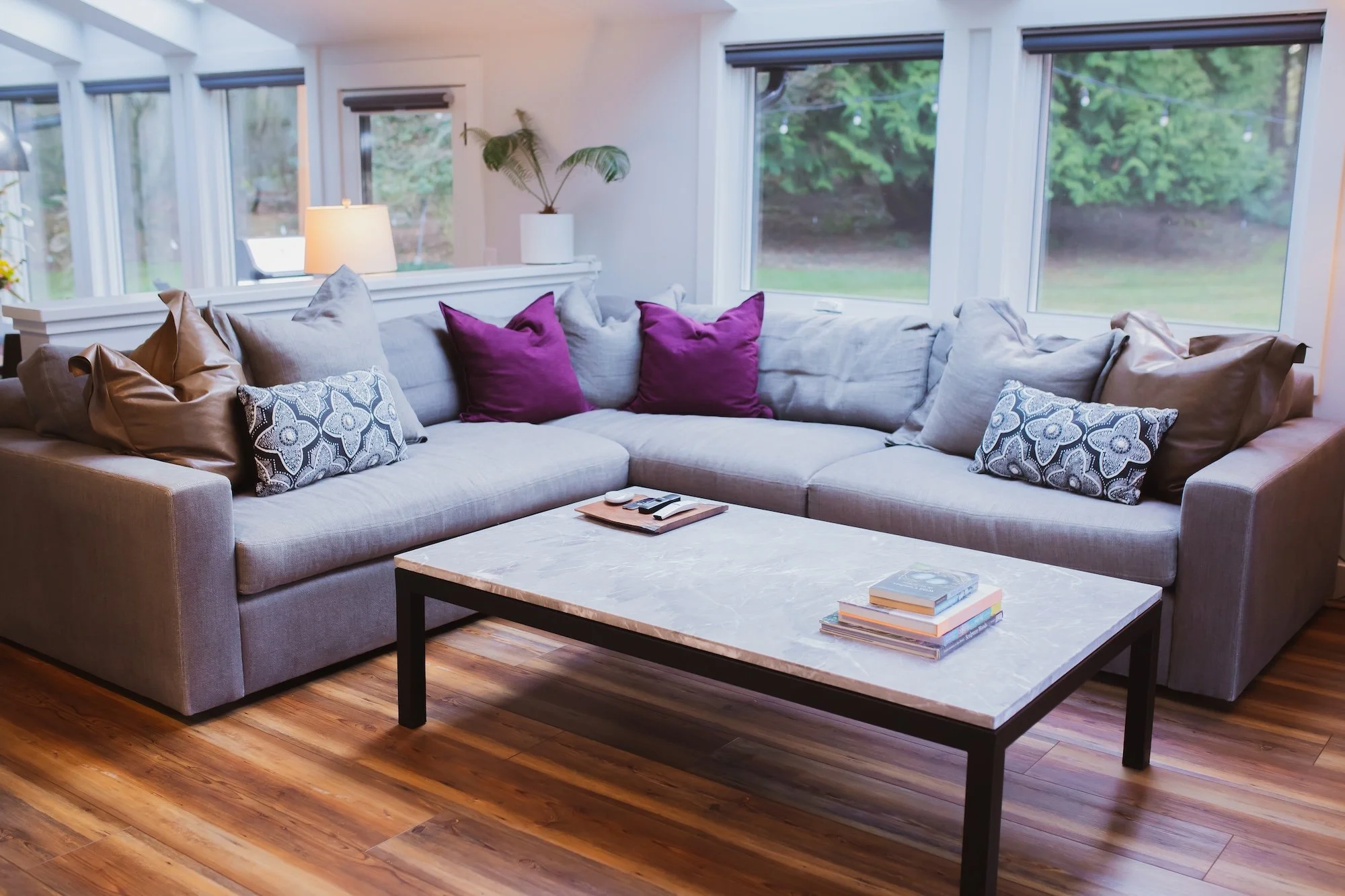 A living room with a large sectional sofa adorned with purple, gray, and patterned pillows, a white marble-top coffee table with books, a lit table lamp, potted plants, and windows showing a view of a green outdoor area.