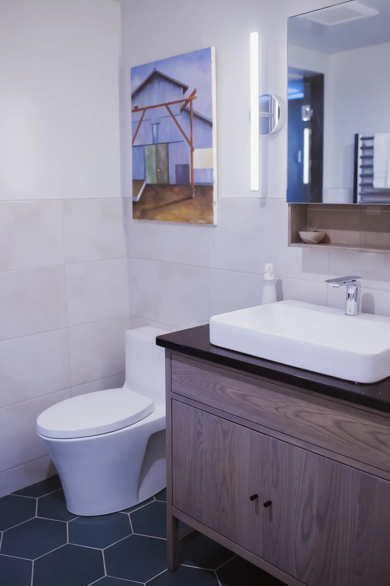 Modern bathroom with white toilet, light wood vanity with black countertop, white sink, wall-mounted mirror, and artwork of an abstract house on the wall.