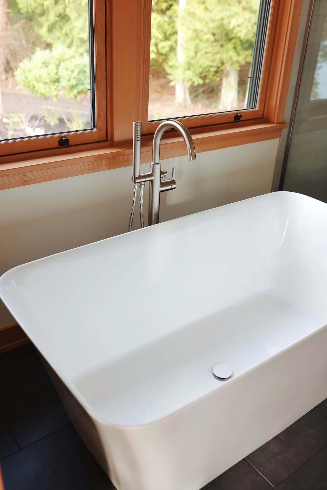 A modern white bathtub with a curved edge, located in a bathroom near two large wooden-framed windows with a view of green trees outside. The bathtub has a sleek silver faucet and a drain at the bottom.