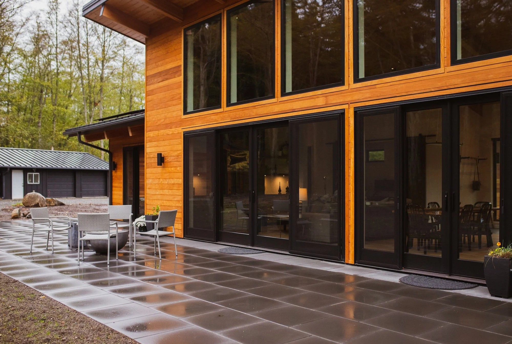 Modern two-story house with large glass windows, wooden exterior, and a patio with outdoor dining furniture, surrounded by trees.