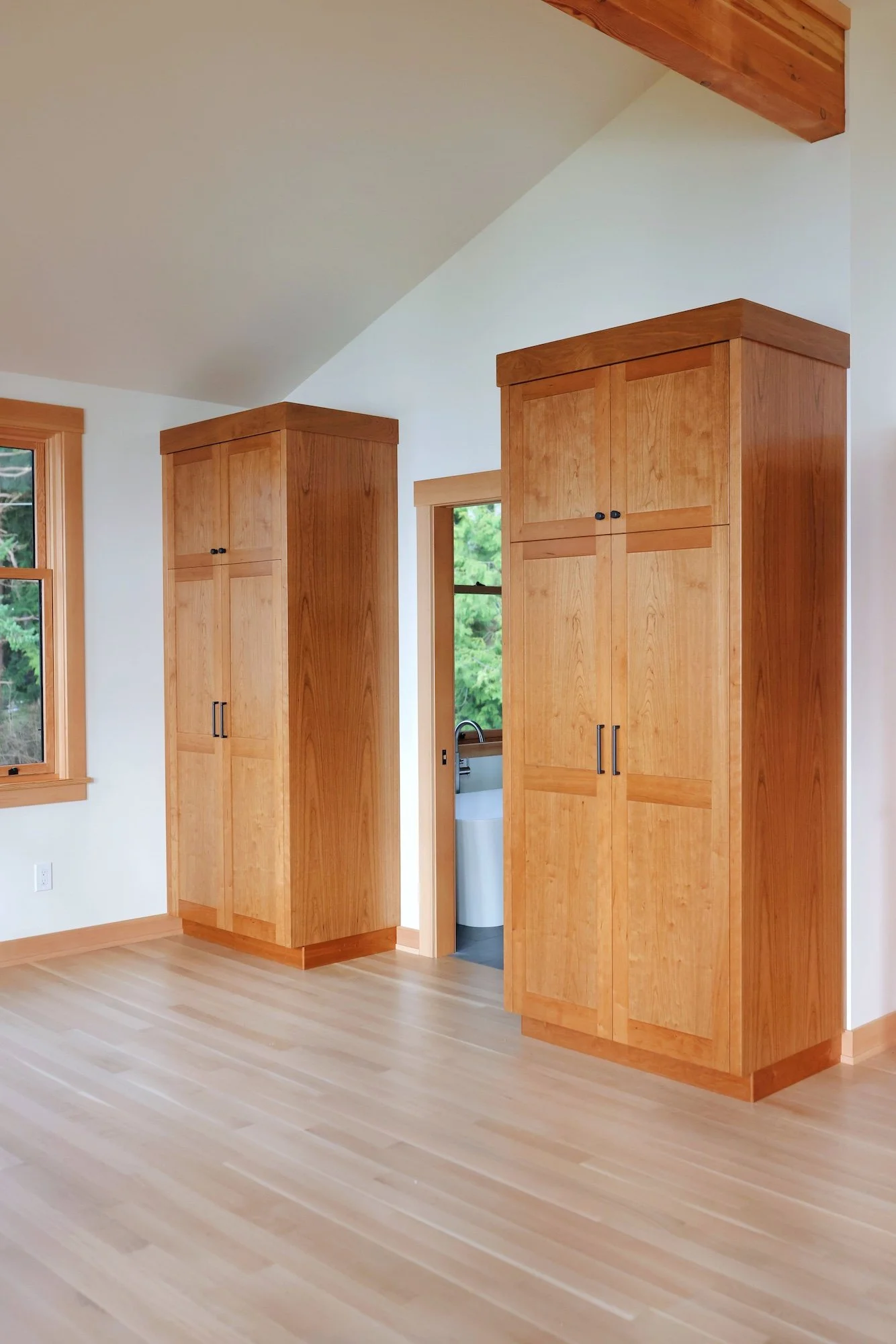 Interior of a room with wooden cabinets, a window, and a sloped ceiling with a wooden beam. The floor is light-colored wood.