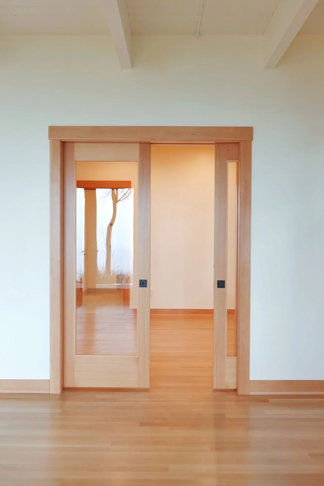 A doorway with natural wood trim opens into a room with wooden flooring and white walls, and a window with a view of a tree outside.