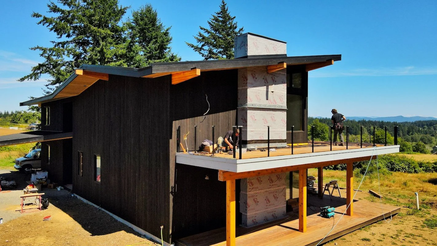 Under construction modern house with black exterior walls and wooden deck, workers installing roofing, in a green open landscape under a blue sky.