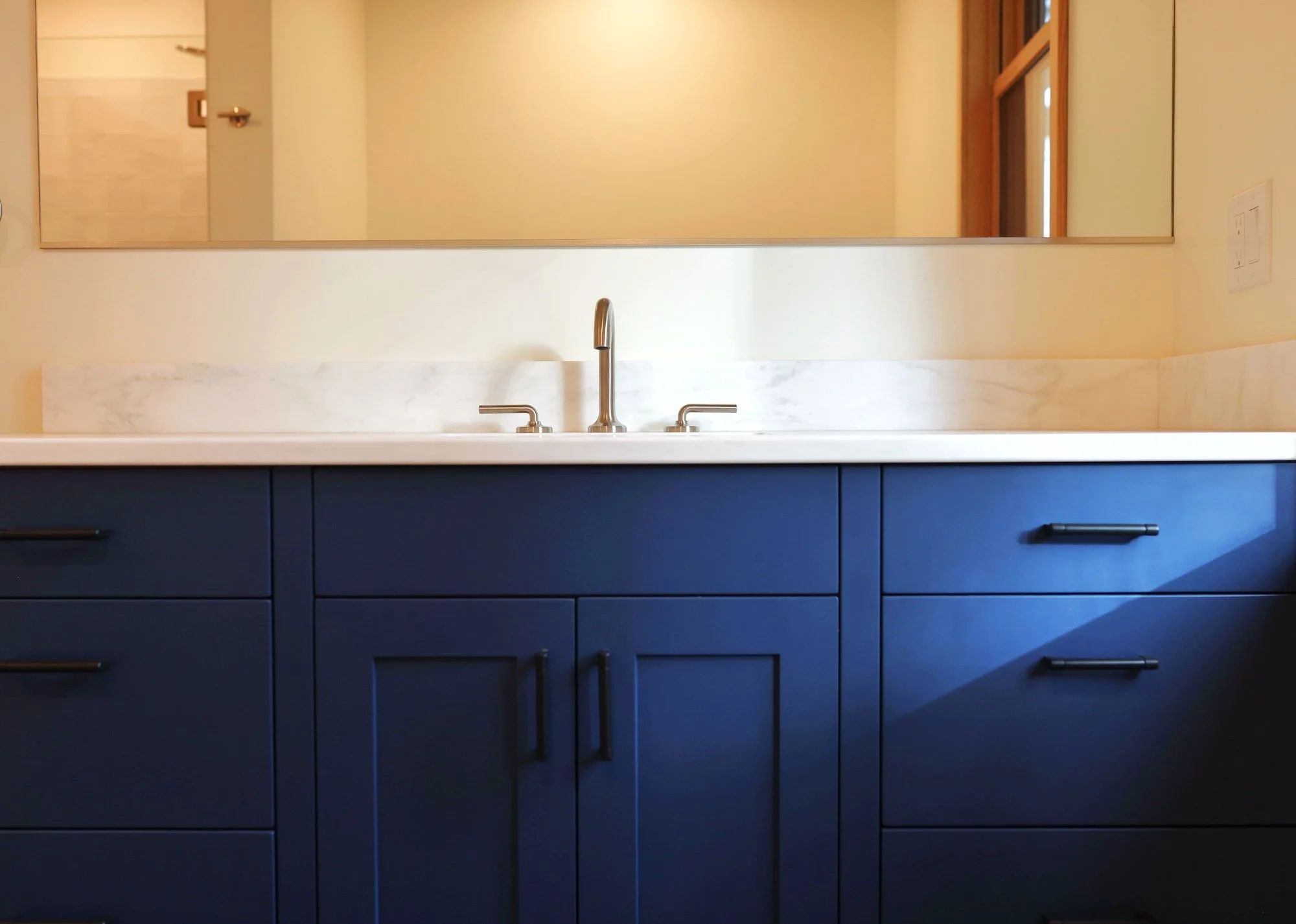 A bathroom vanity with dark blue cabinets, a white marble countertop, and a brass-colored faucet. A mirror above the sink reflects part of the room, including a door and a window.