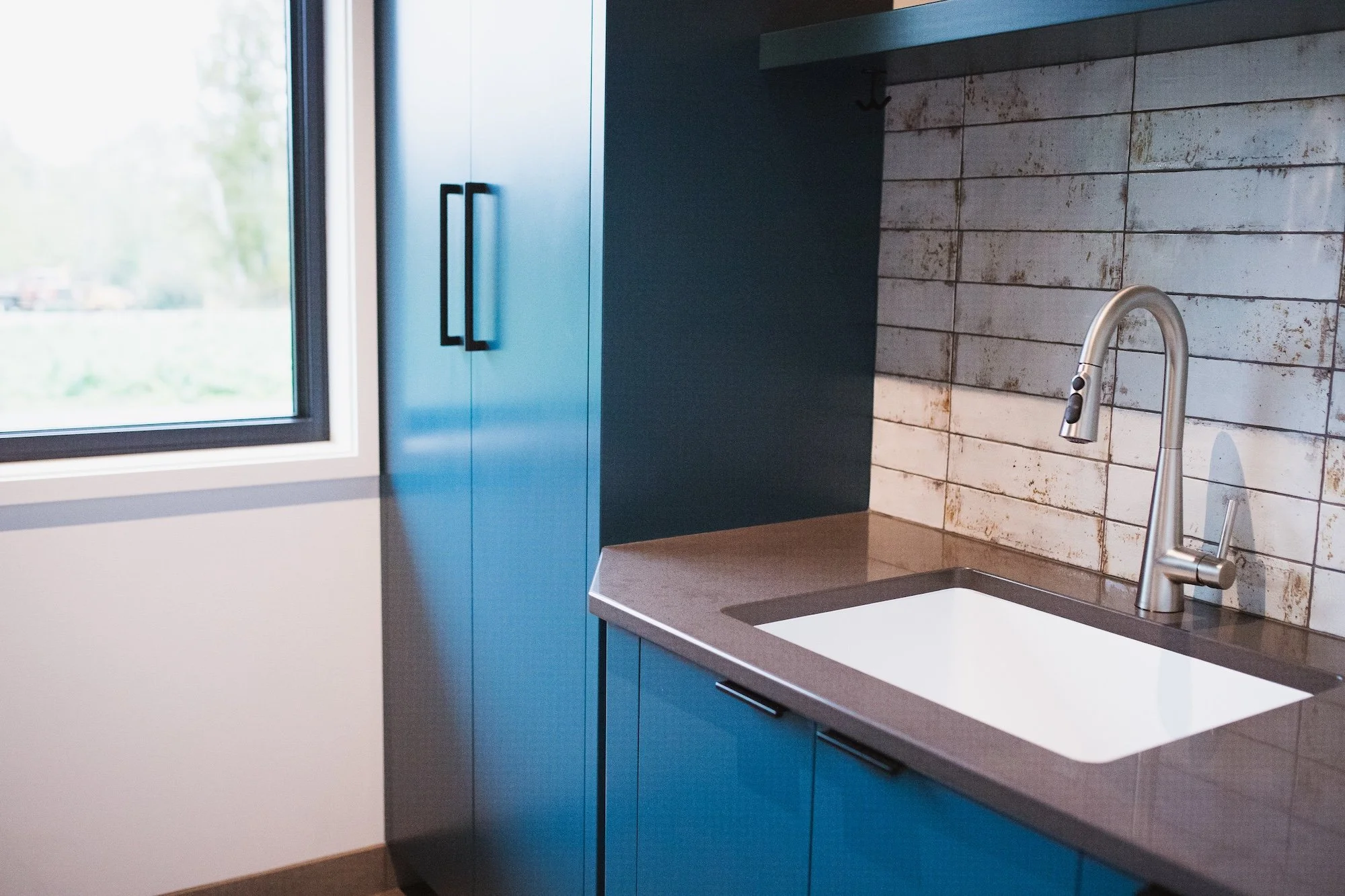Modern kitchen with blue cabinets, a white undermount sink, and a silver faucet, with a window showing an outdoor view and a tiled backsplash.