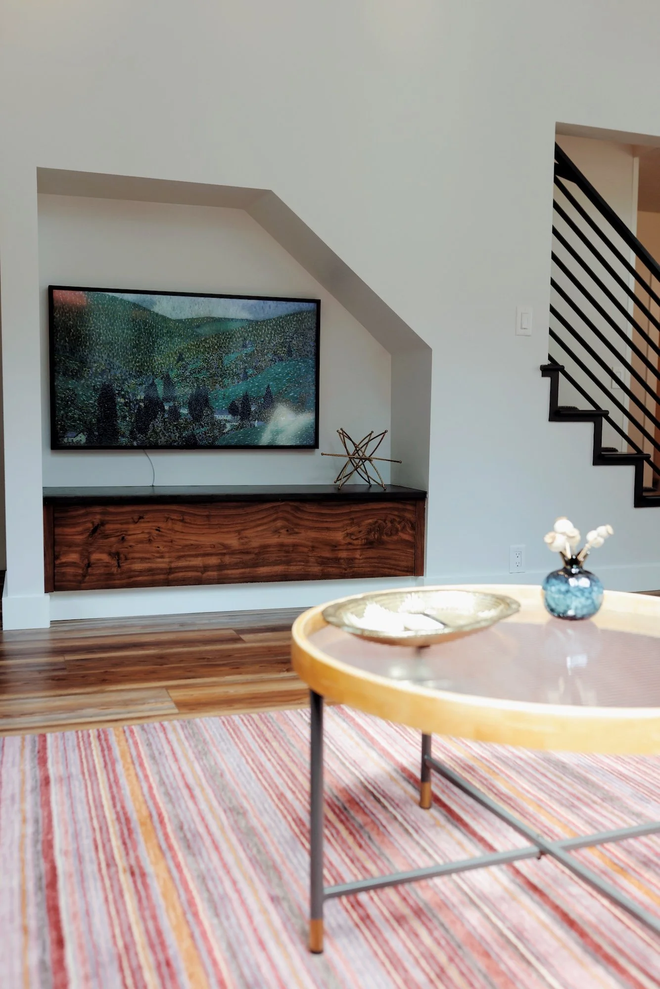 Living room with a mounted television, a wooden console, a decorative geometric sculpture, a round wooden table, and a colorful striped rug.