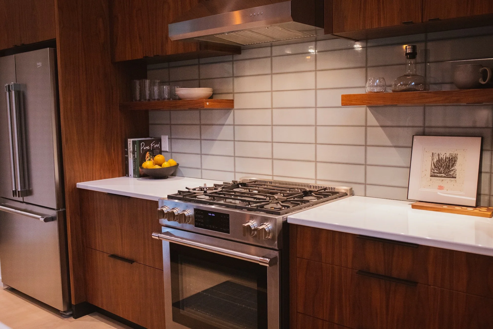 Modern kitchen featuring a stainless steel refrigerator, a gas stove, wooden cabinets, open shelves with glassware and bowls, a white countertop with a bowl of lemons, and framed artwork leaning against the backsplash.