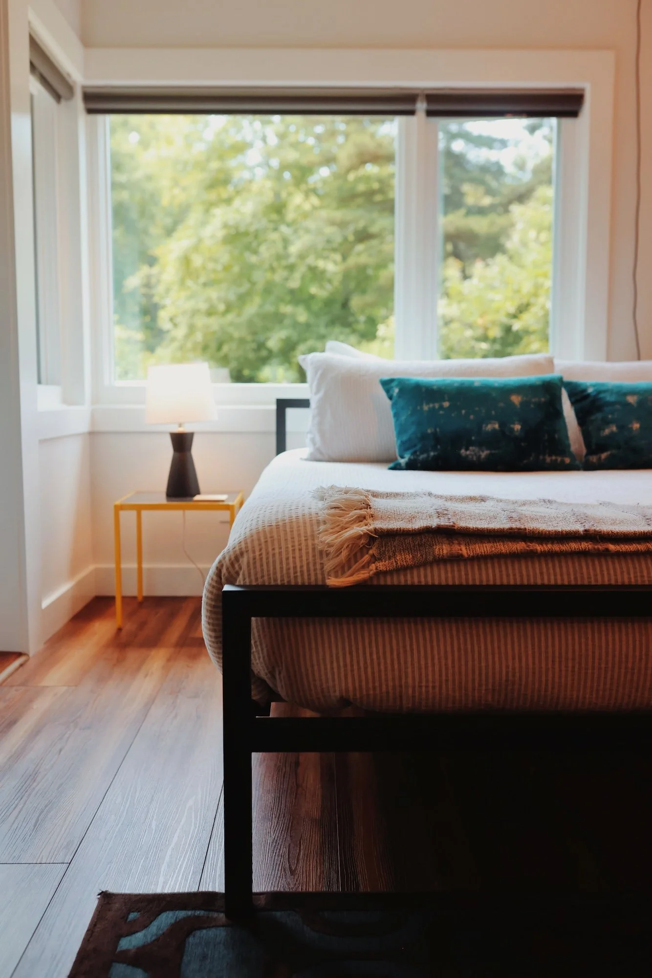 A bedroom with a large window showing green trees outside. There is a bed with white and blue velvet pillows, a beige blanket, and a wooden floor. A small yellow table with a black lamp and a white lampshade is next to the bed. A brown patterned rug 