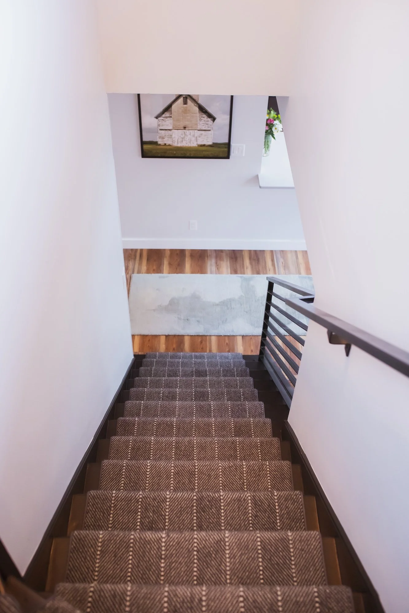 View down a staircase with patterned carpeted steps, white walls, a black and white photograph of an old building on the wall, a vase with flowers on a ledge, and hardwood flooring at the bottom.