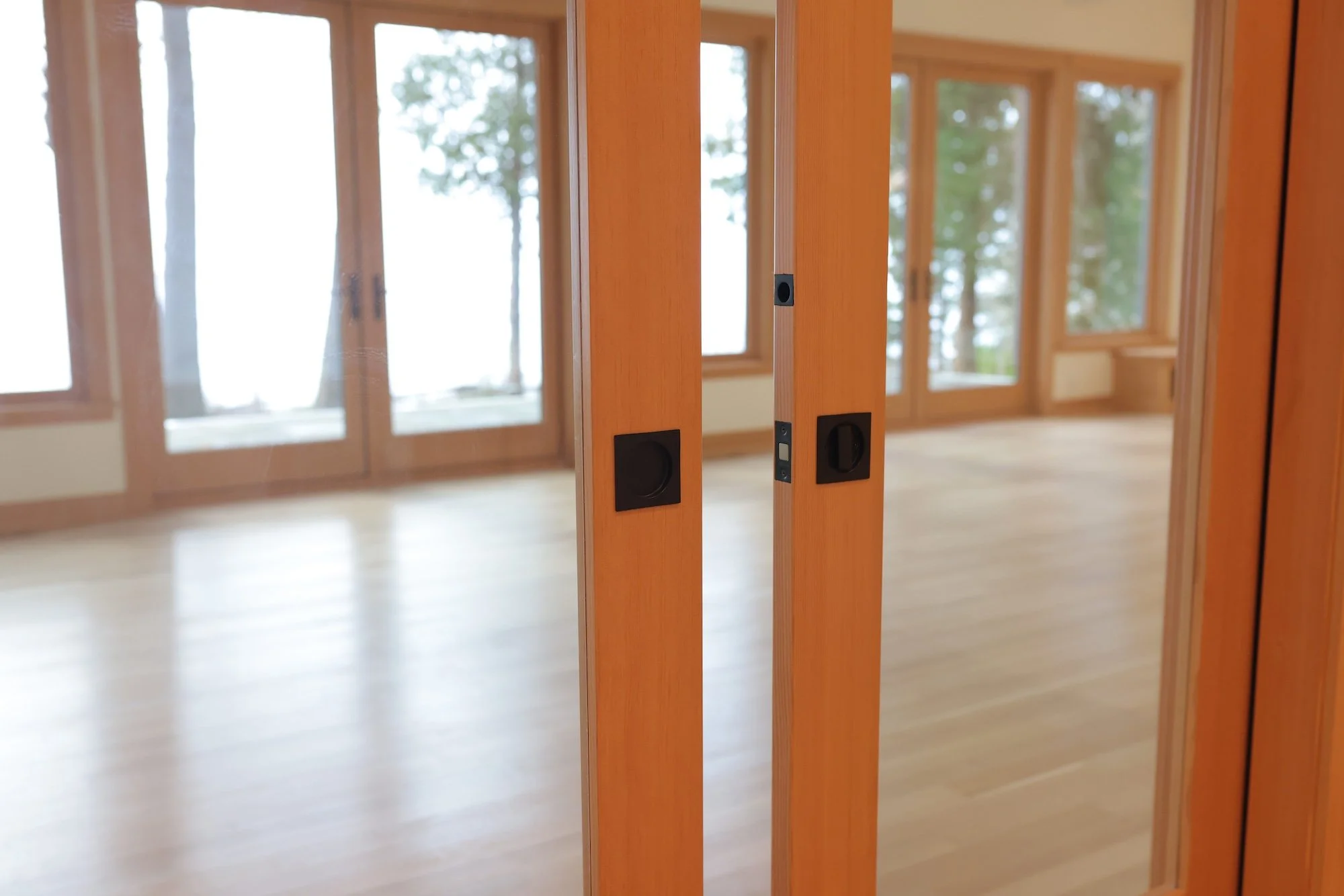 Close-up of a wooden French door frame with a black door knob, opening into a room with wooden floors and large windows showing trees outside.
