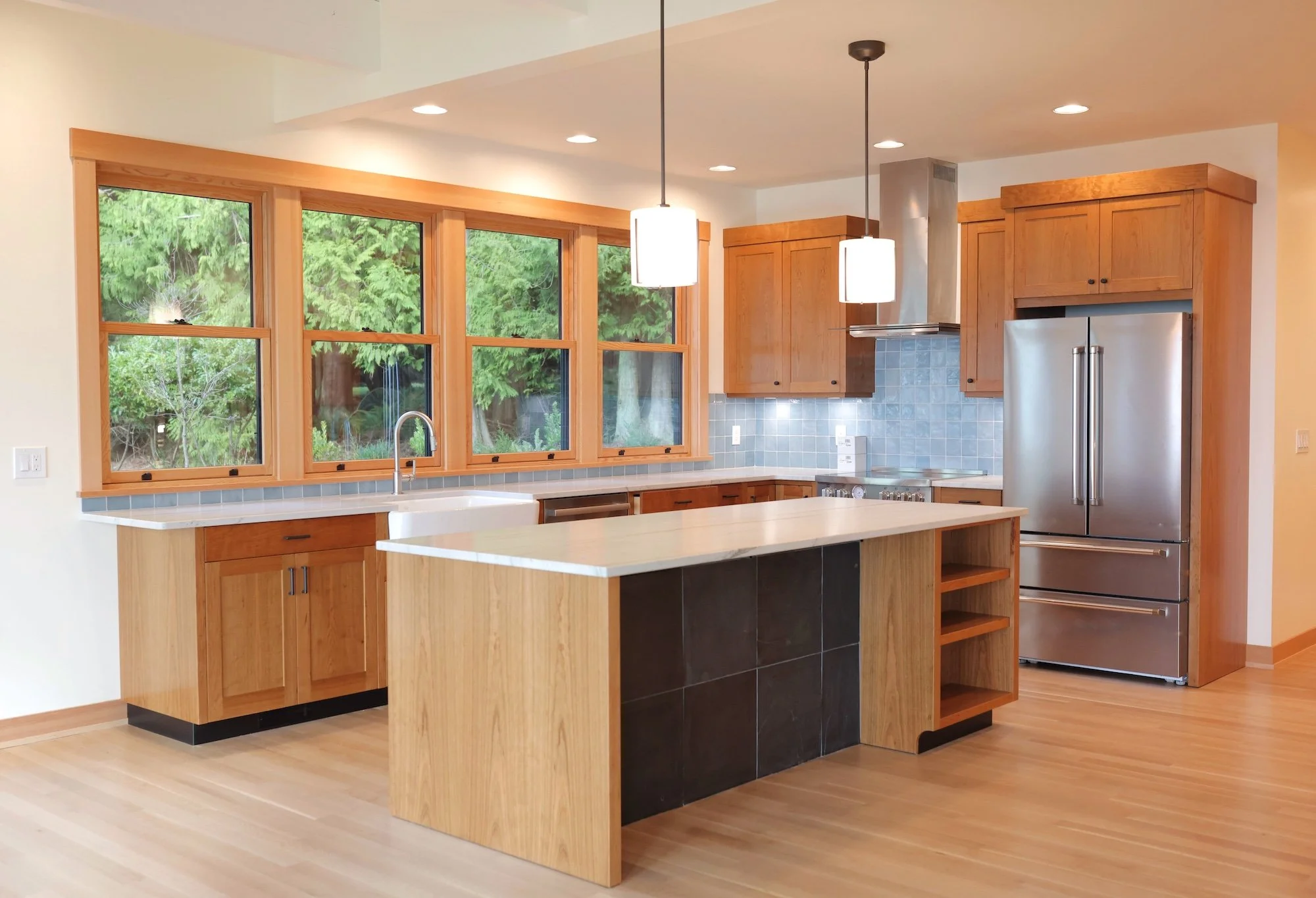 A modern kitchen with large windows, wooden cabinets, a central island with black tile base, hardwood floors, and stainless steel appliances, including a refrigerator and range hood. Pendant lights hang above the island.