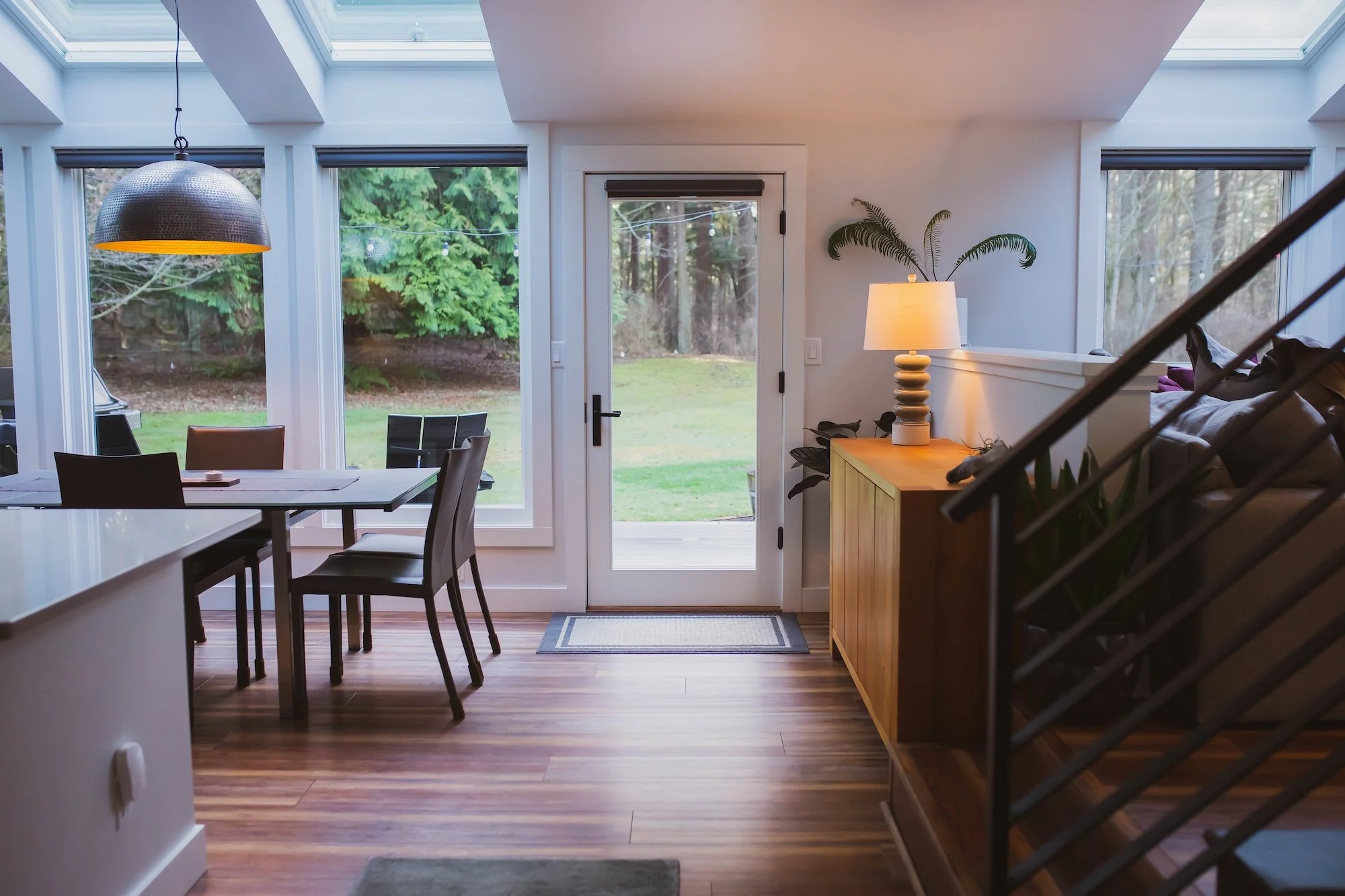 A bright room with large windows and skylights, a dining table with chairs, a door leading outside onto a grassy yard, a wooden sideboard with a lamp and houseplants, and a staircase partially visible on the right.
