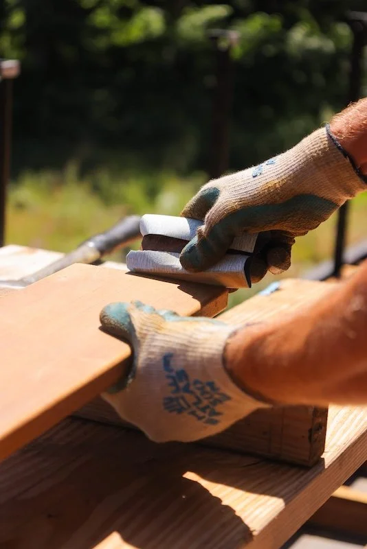 Hands wearing gloves are smoothing a piece of wood with a tool outdoors in bright sunlight, with green trees in the background.