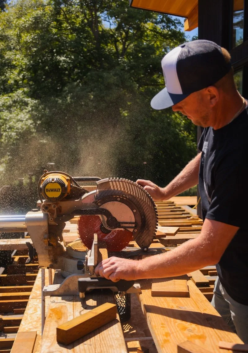 A man wearing a baseball cap is using a DeWalt miter saw to cut a piece of wood outdoors during daytime.