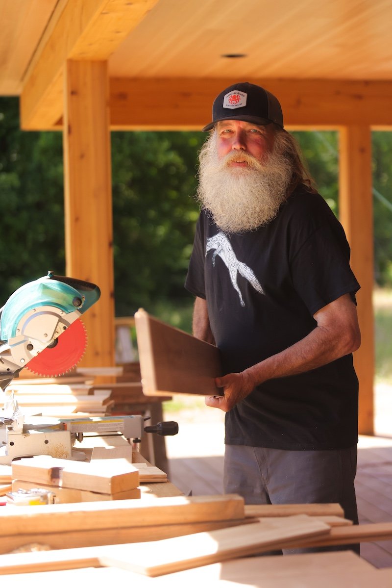 An older man with a long white beard and mustache, wearing a black baseball cap and black t-shirt, is holding a piece of wood with a miter saw and a workbench in front of him, in an outdoor woodworking shop with a wooden roof and green trees in the background.