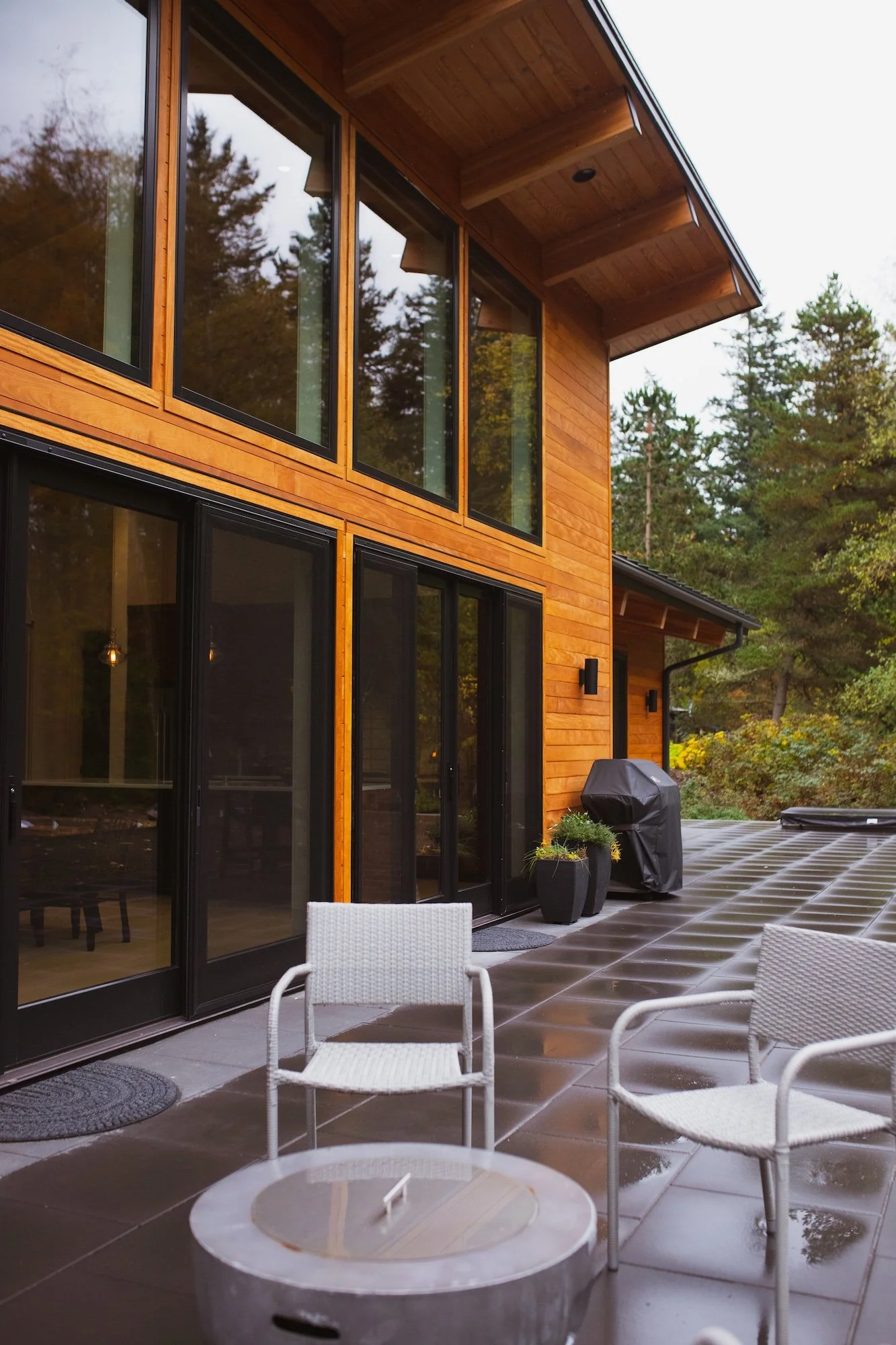 A modern house with large glass windows and a wooden exterior, surrounded by trees. There is an outdoor patio with chairs and a table, and some plants and a barbecue grill.