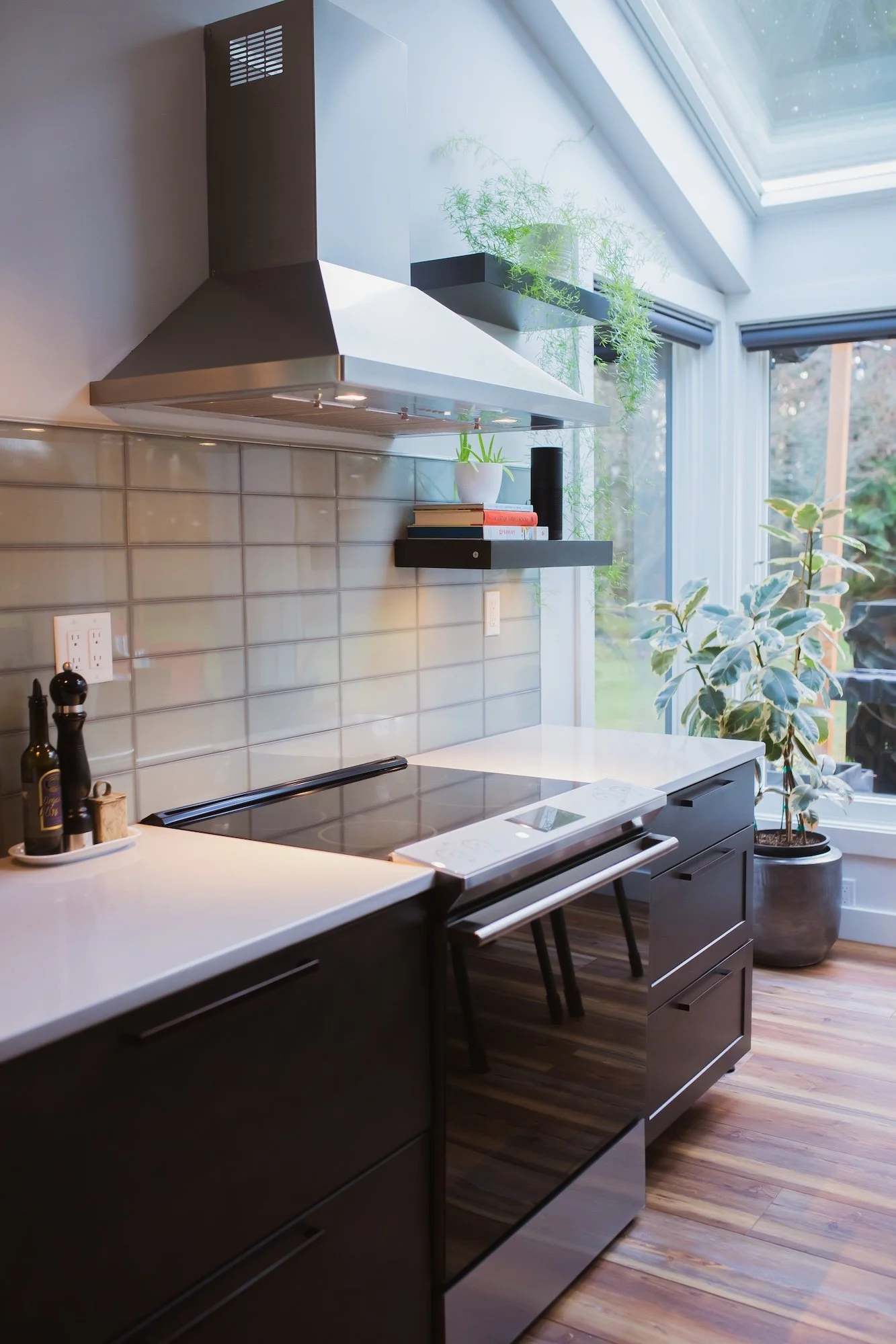 A modern kitchen with white countertops, dark cabinets, a stainless steel stove and hood, a tiled backsplash, and large windows with a view of a garden outside. There are potted plants and a few cookbooks on a black shelf above the counter.