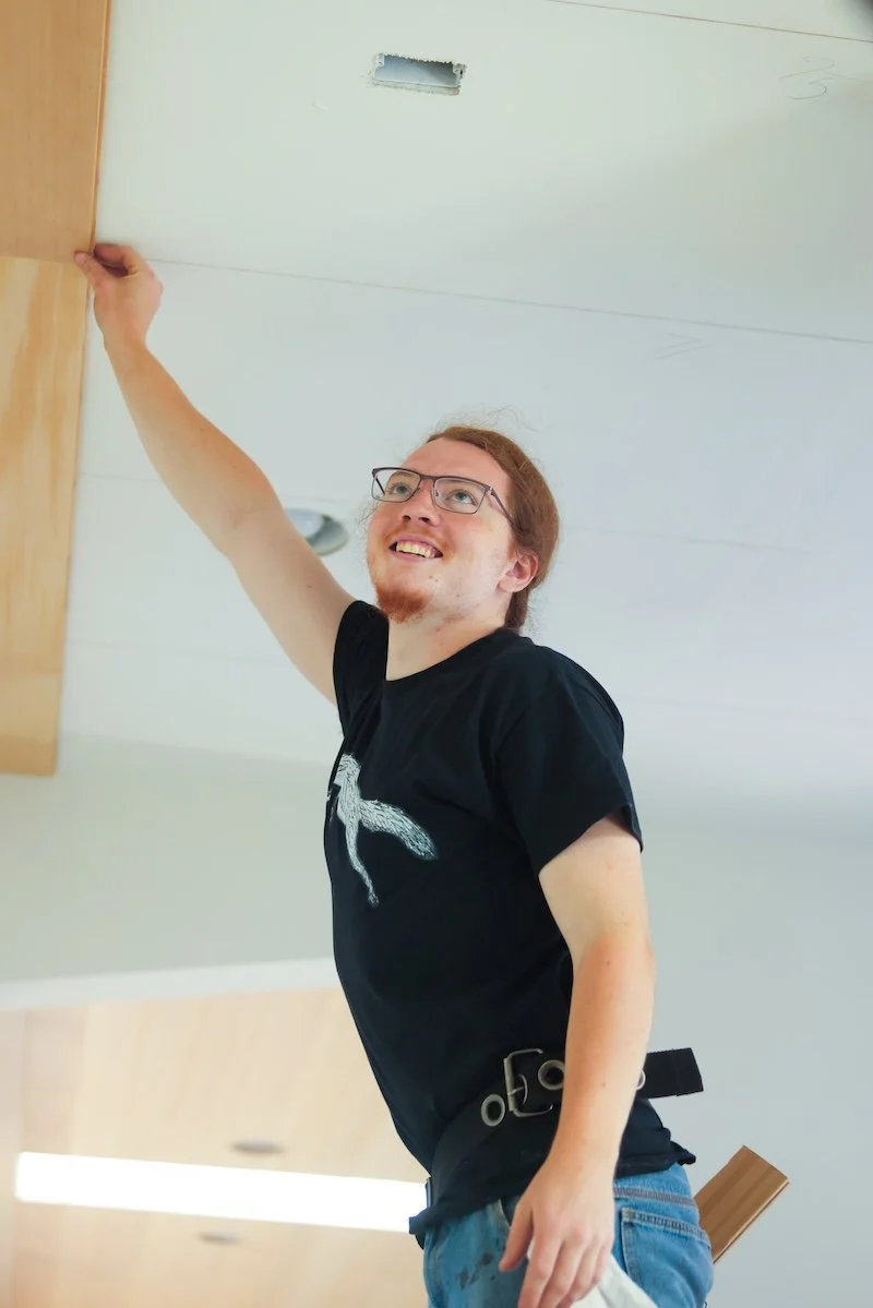A man with glasses and red hair smiling as he installs or repairs a wooden cabinet near the ceiling.