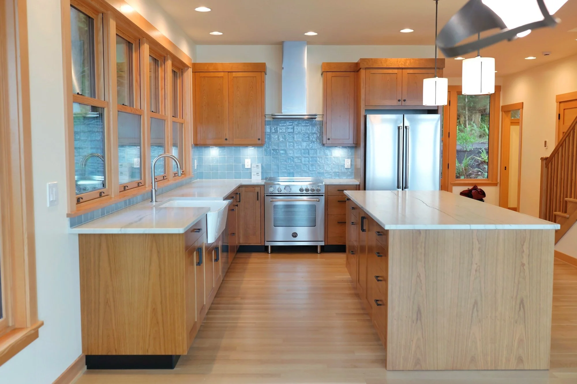 A modern kitchen with wooden cabinets, a stainless steel refrigerator, and a stove with a hood, illuminated by ceiling lights and pendant lamps, with windows showing outdoor greenery.