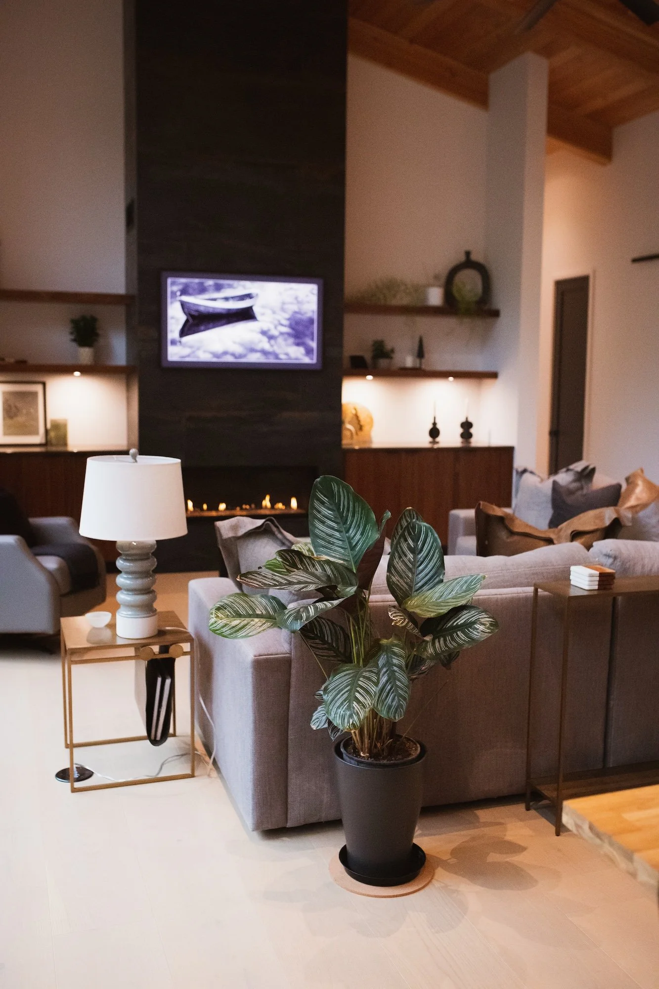 A cozy living room with a beige sectional sofa, a potted plant, a white table lamp, and wooden shelves with decorative items. A wall-mounted TV displays a black-and-white photo of a boat, and ambient lighting creates a warm atmosphere.