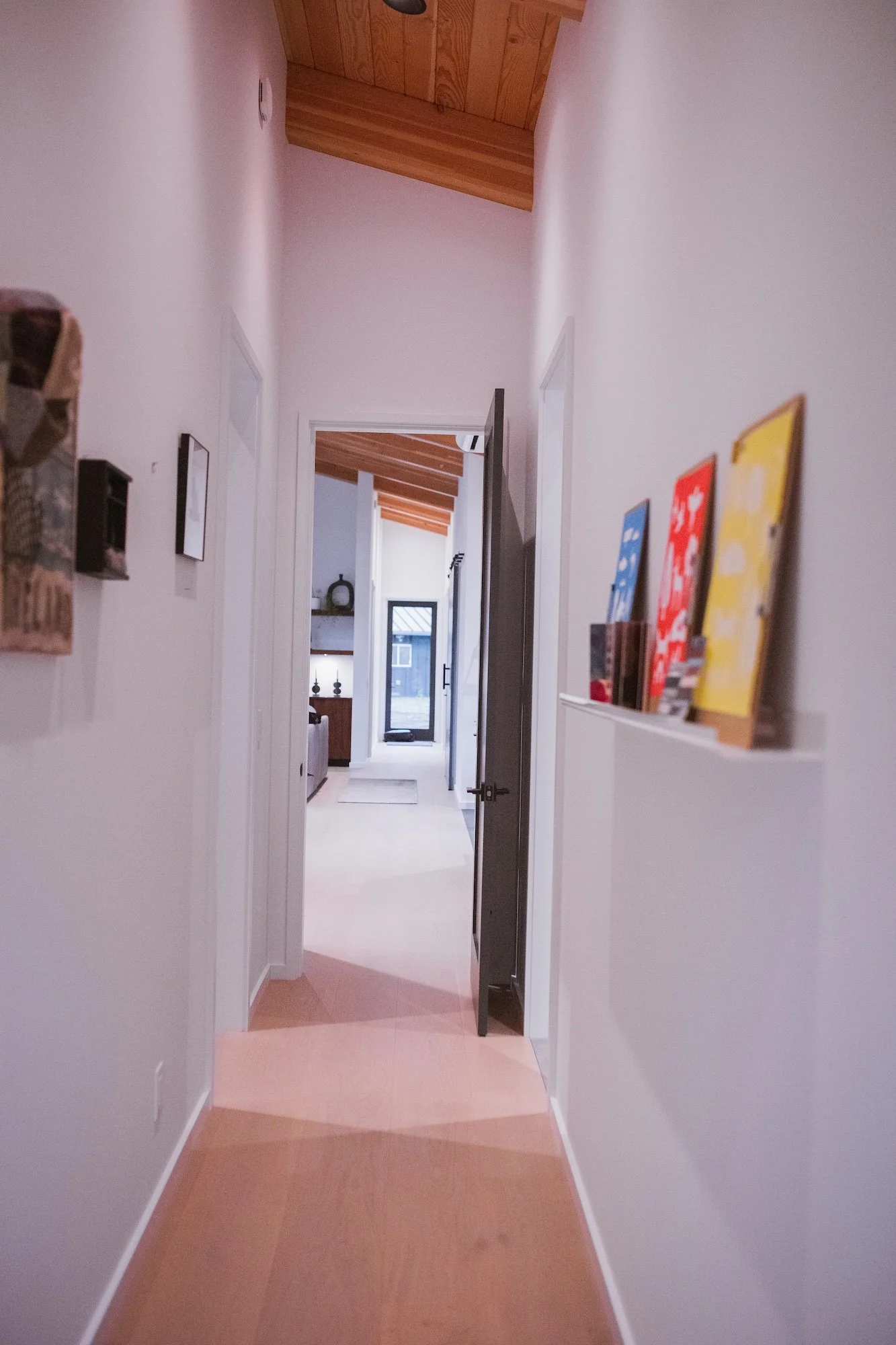 View of a hallway leading to a bright room with a glass door and a small outdoor area visible outside. The hallway has light pink walls, wooden ceiling, and a beige tile floor. Artwork is displayed on the walls.