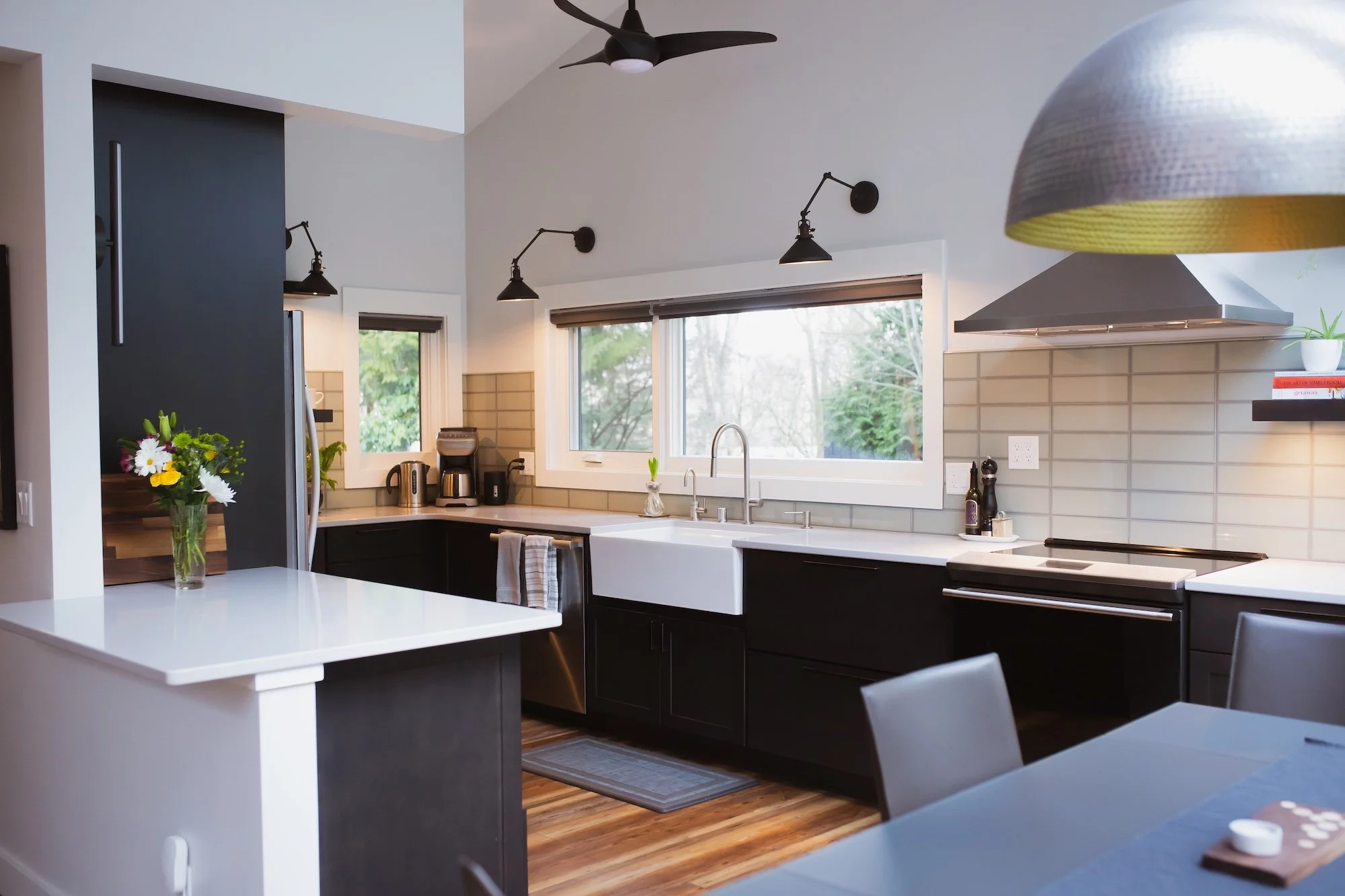 Modern kitchen with black cabinets, white countertops, stainless steel appliances, and large windows overlooking trees, with black wall-mounted lamps and a ceiling fan.
