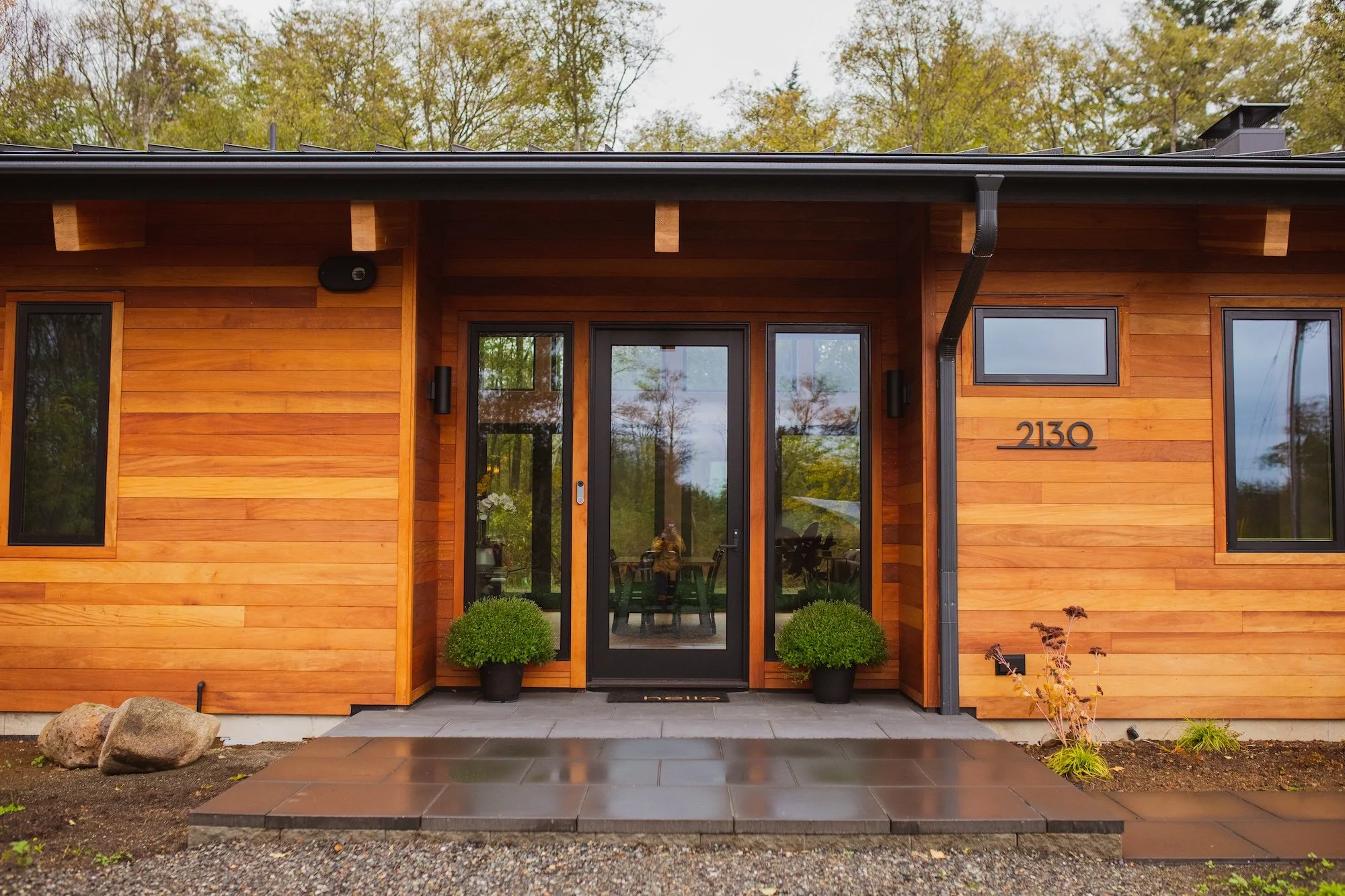 Front view of a modern house with wooden exterior, black front door with glass panels, and two potted plants on each side of the door. There are two small windows on each side of the door, a black gutter running down the right side, and mature trees 