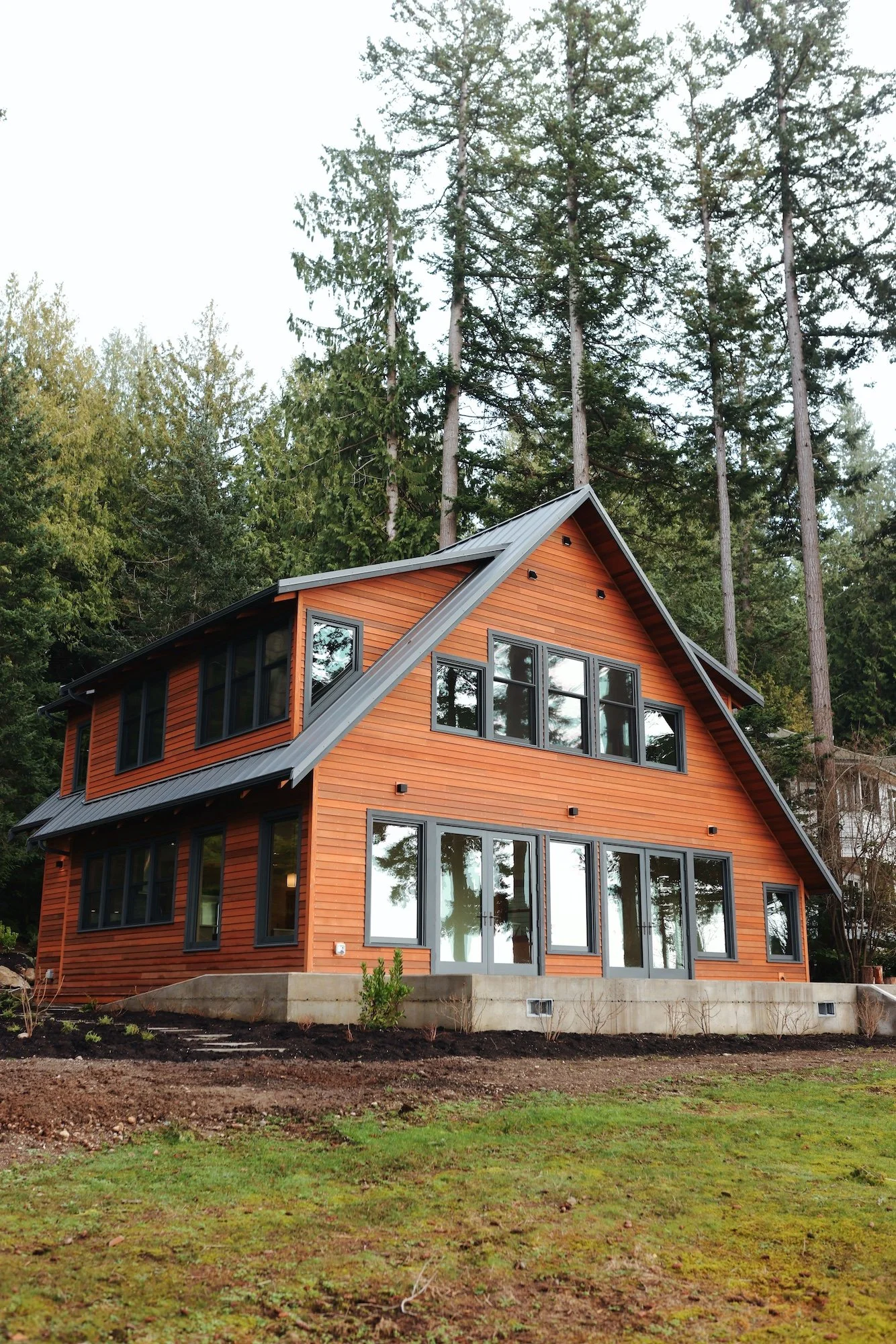 A modern, multi-story house with large windows and a metal roof, built on a concrete foundation, surrounded by grass and trees, with tall pine trees in the background.