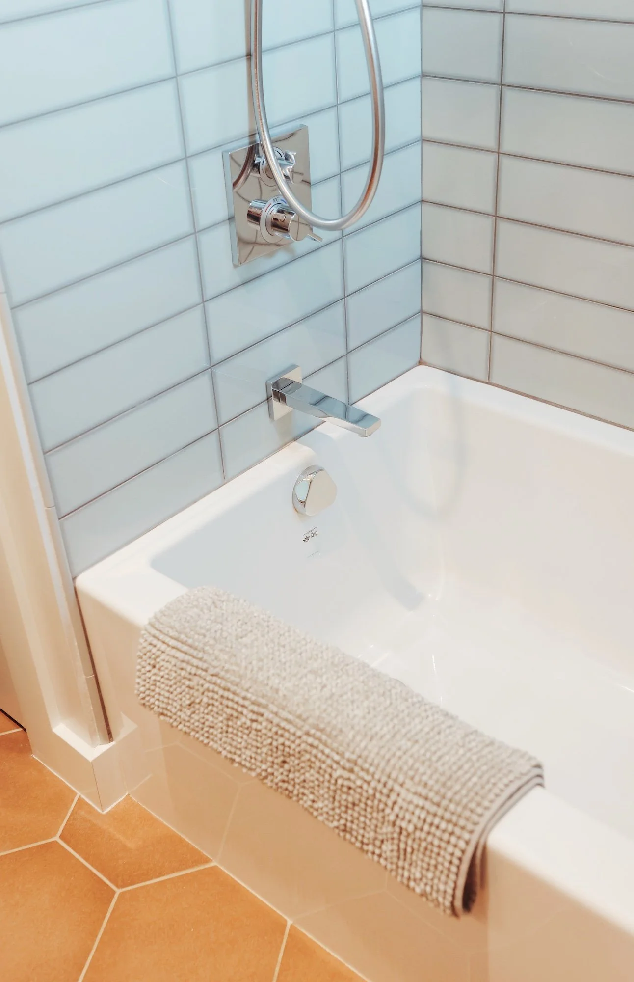 A bathroom bathtub with a beige towel hanging over the edge, white tiled walls, and a chrome faucet with a hand shower.