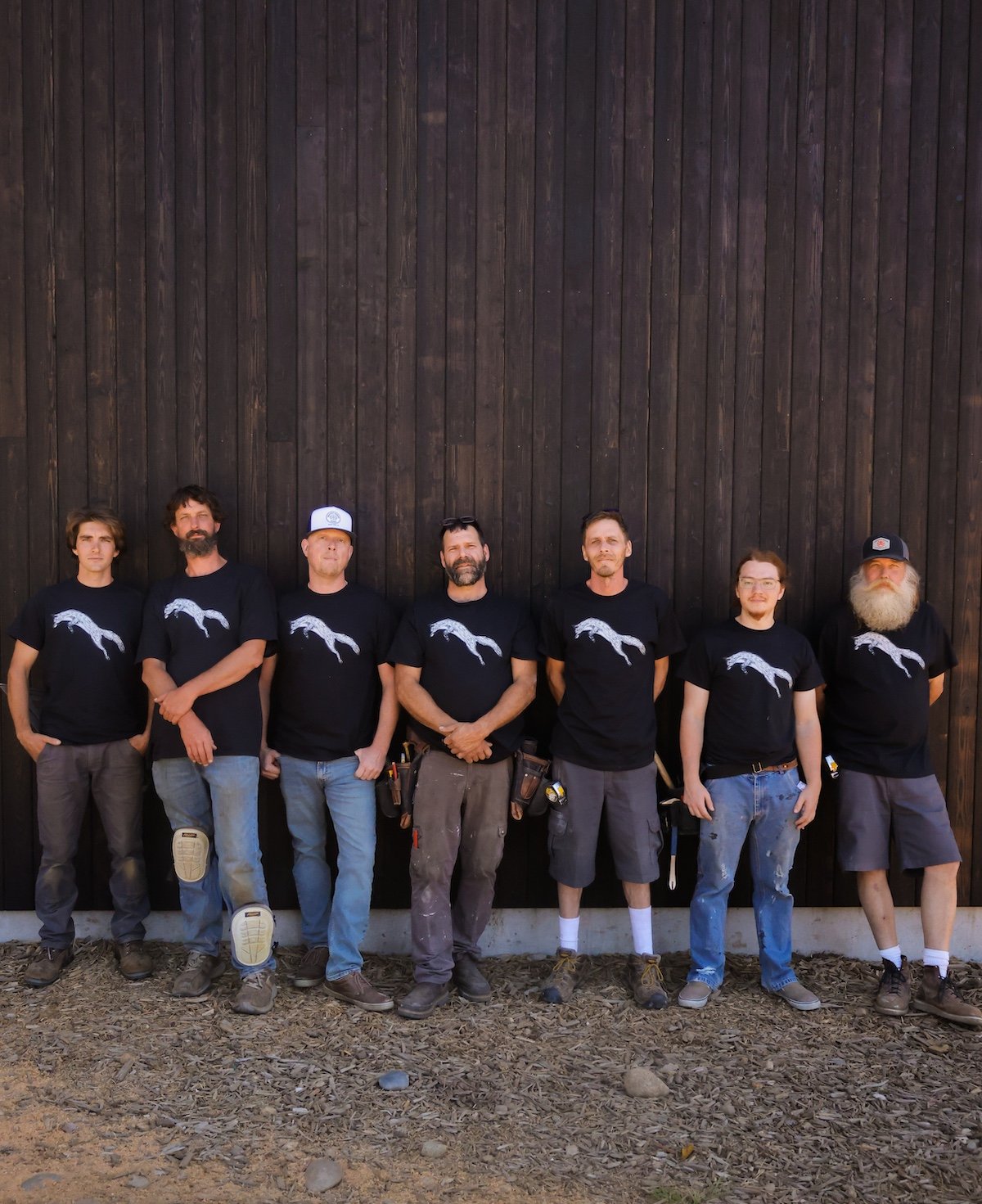 A group of seven men standing side by side in front of a large wooden wall. They are wearing black T-shirts with a white graphic of a horse on the front, and casual pants or shorts. They have varying hairstyles and facial hair, with some wearing hats or glasses. Some have work tools or equipment attached to their belts.