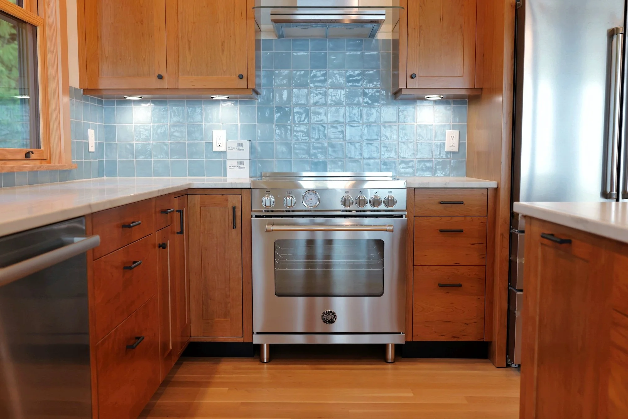 A kitchen with wooden cabinets, light blue tiled backsplash, stainless steel stove, and a window on the left side.