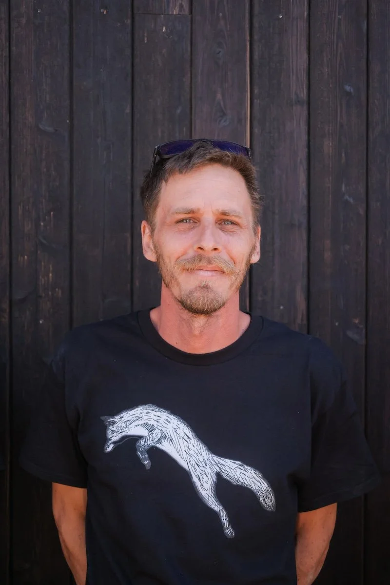 A man with short brown hair, a beard, and light skin, standing outdoors in front of a dark wooden fence. He is wearing sunglasses on his head and a black T-shirt with a white sketch of a jumping fox on it.