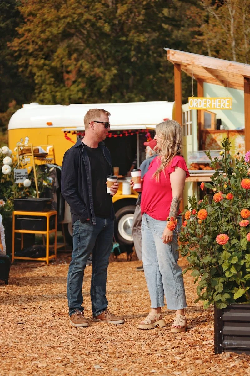 Two people, a man and a woman, are standing and talking at a food truck event during autumn, both holding coffee cups. The woman has tattoos and is wearing a red shirt and jeans, while the man is wearing glasses, a black shirt, and a dark jacket. In the background, there is a yellow food truck and a sign that says "Order Here!" with fall foliage around.