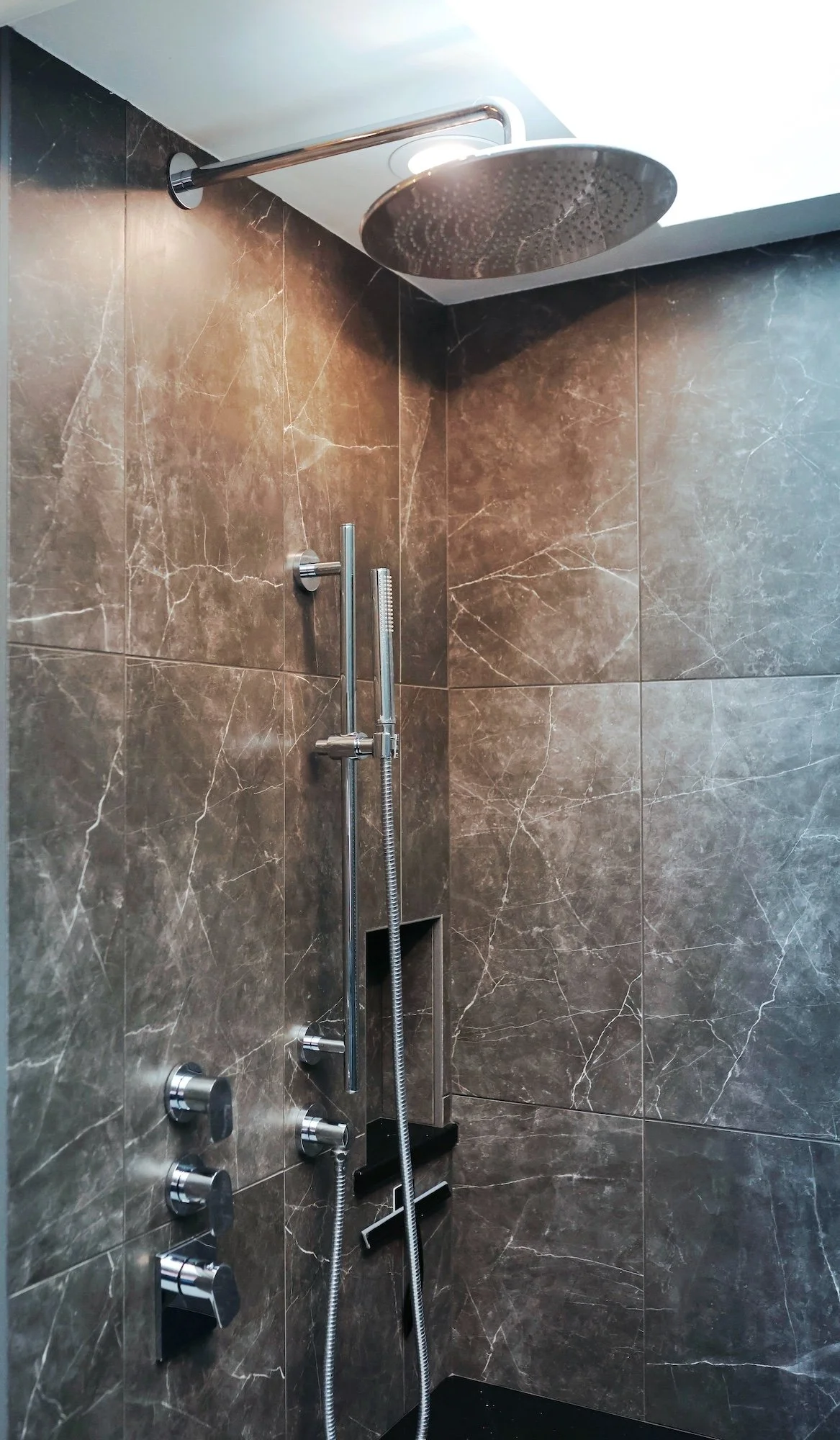 A modern shower with a large round rain shower head, a handheld shower wand, and multiple control knobs on a dark marble wall.