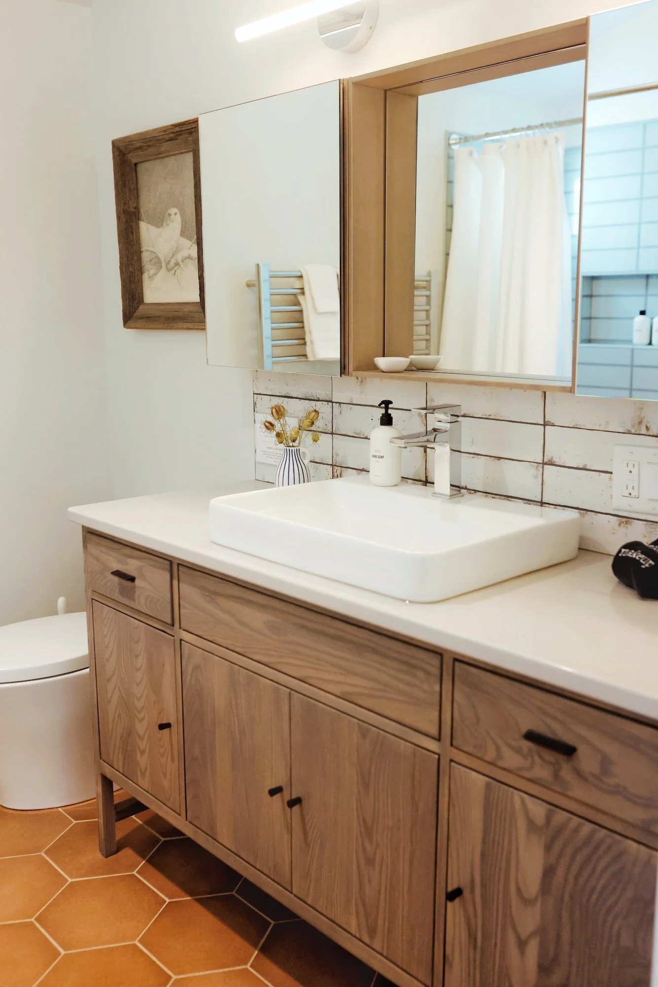 Bathroom vanity with a white sink, wooden cabinets, and a silver faucet. A large mirror is above the sink, with a shelf holding small bowls. Beyond the mirror, a shower with white curtains and a metallic towel rack with white towels are visible. On t