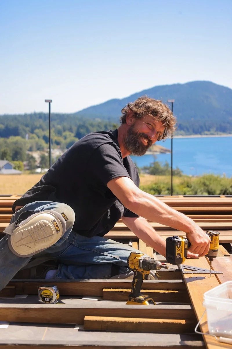 A man with curly hair and a beard installing wood on a deck outdoors with a scenic lake and mountains in the background, using power tools.