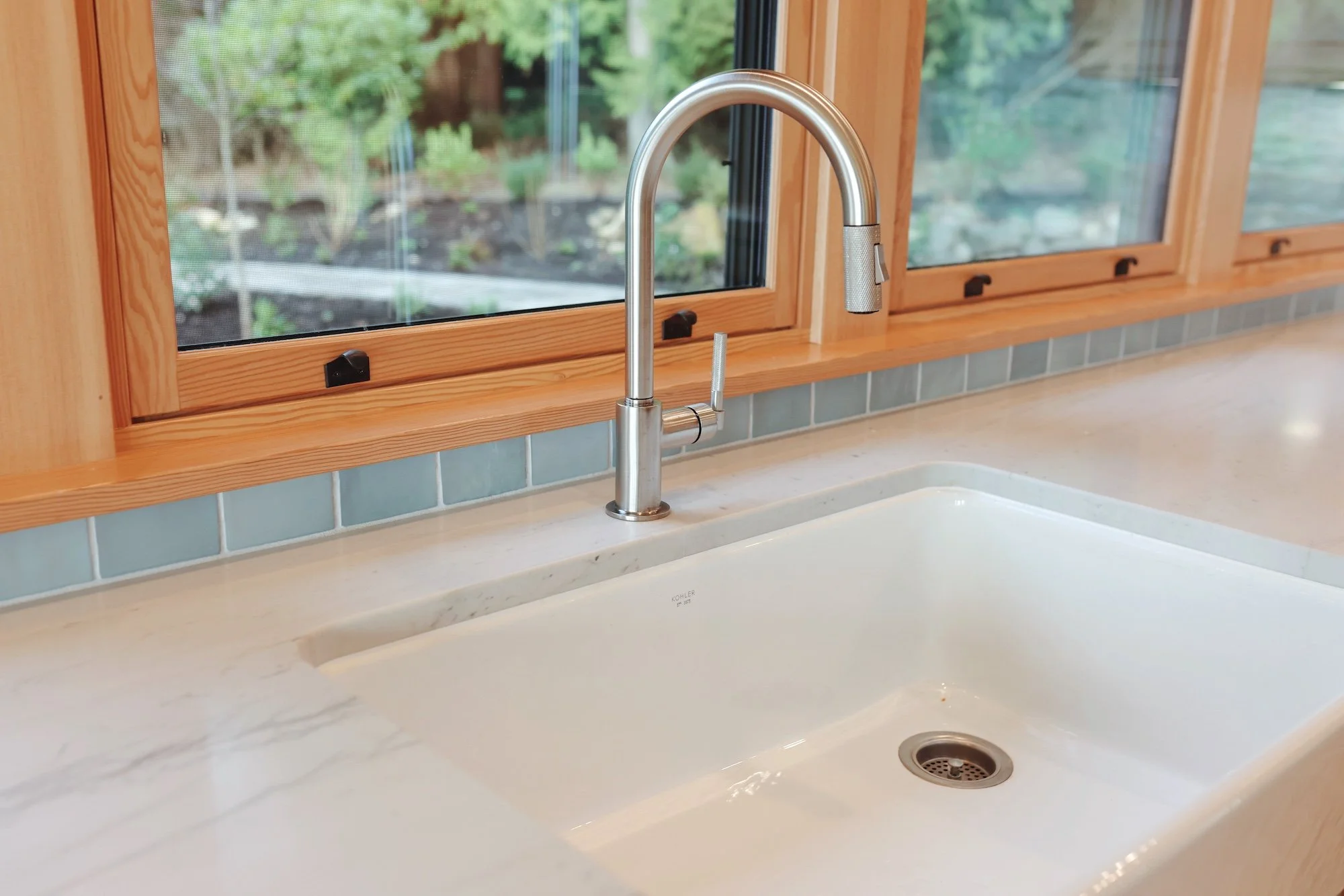A kitchen sink with a modern silver faucet, set into a white marble countertop. Behind the sink is a large window with a wood frame, showing a view of a garden with green plants and trees.