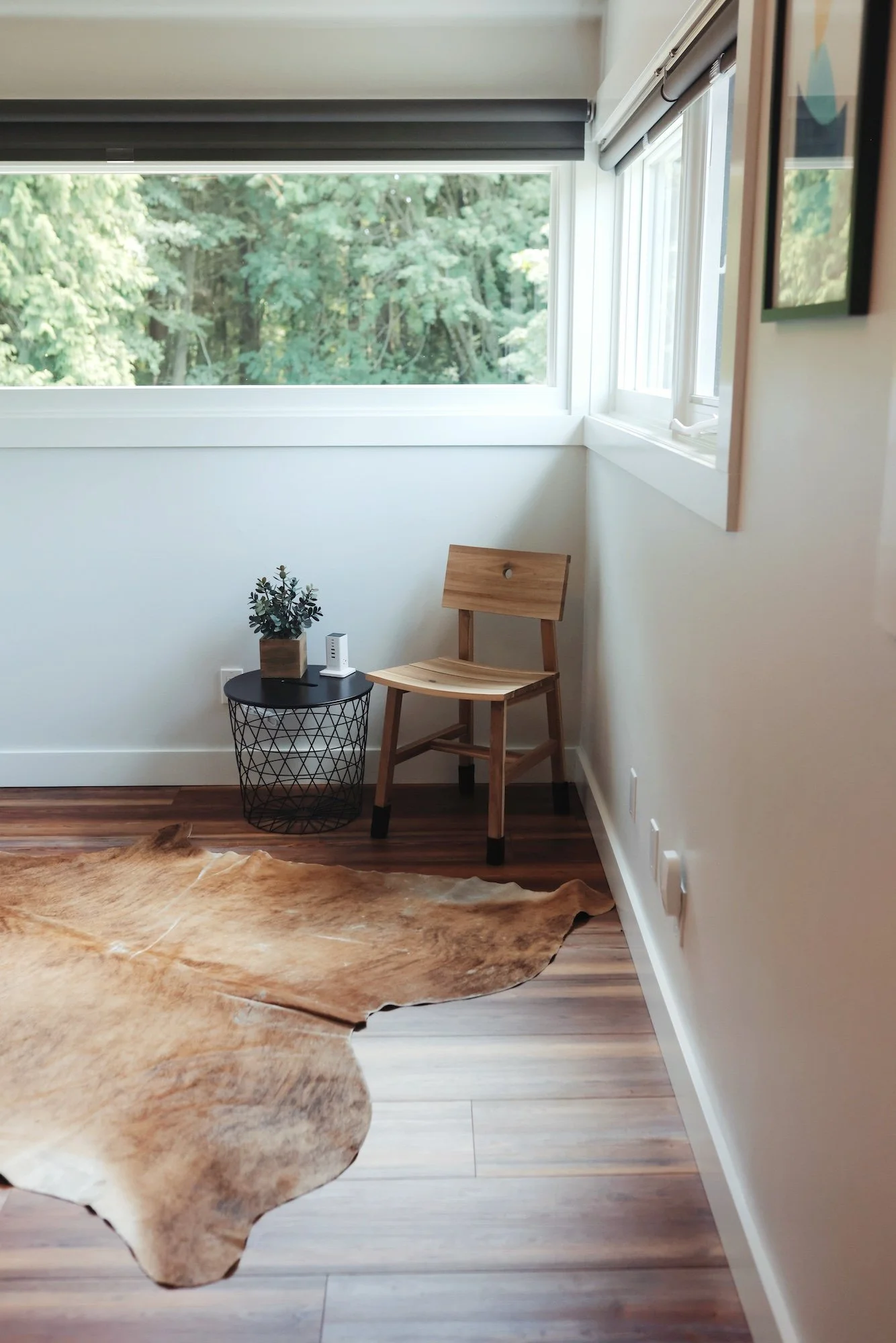 A corner of a room with a large window and a view of trees outside. There is a wooden chair, a small black wire side table with a potted plant and modem, and a brown animal hide rug on the hardwood floor.